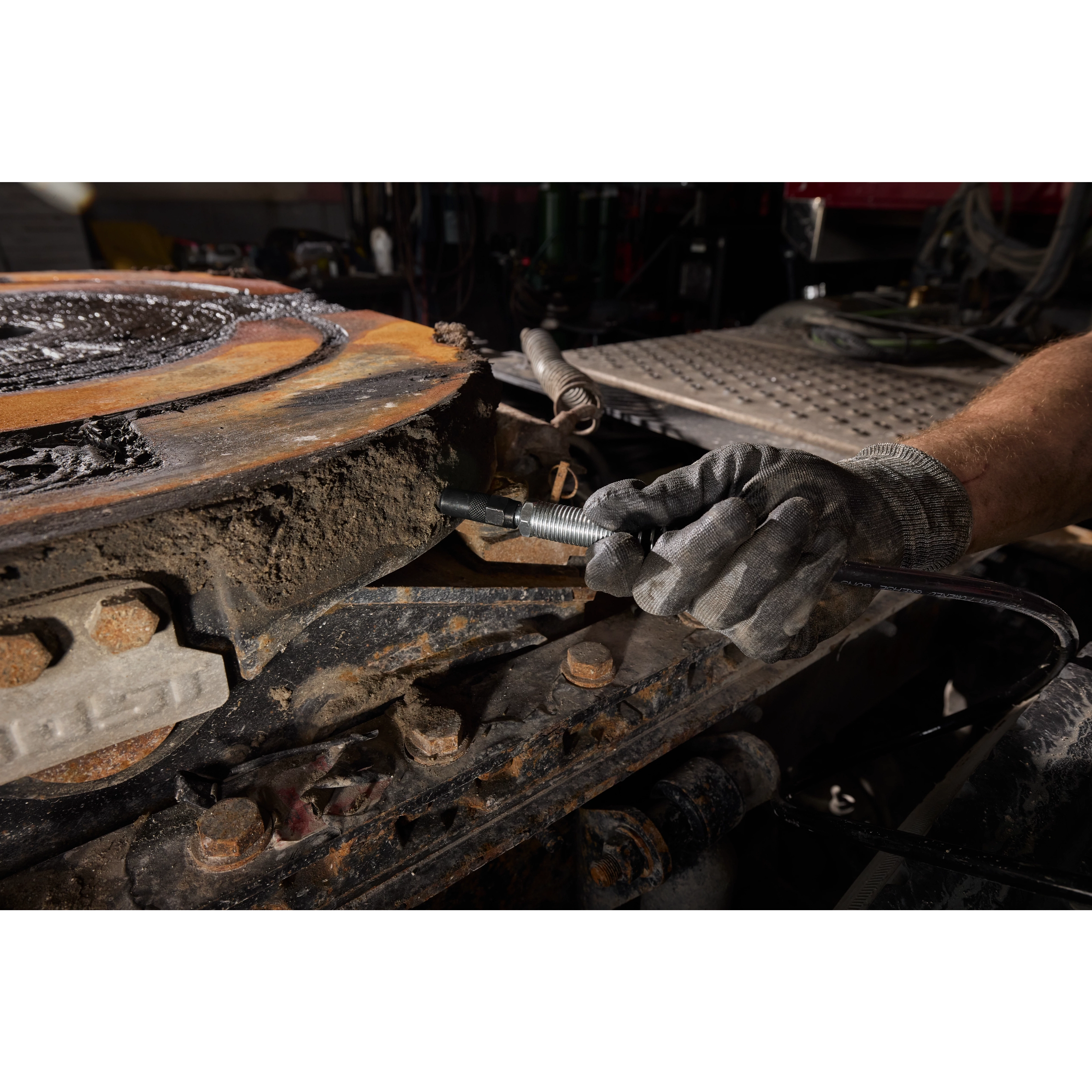Gloved hand using a Right Angle Grease Coupler on a dirty, rusted metal machinery part.