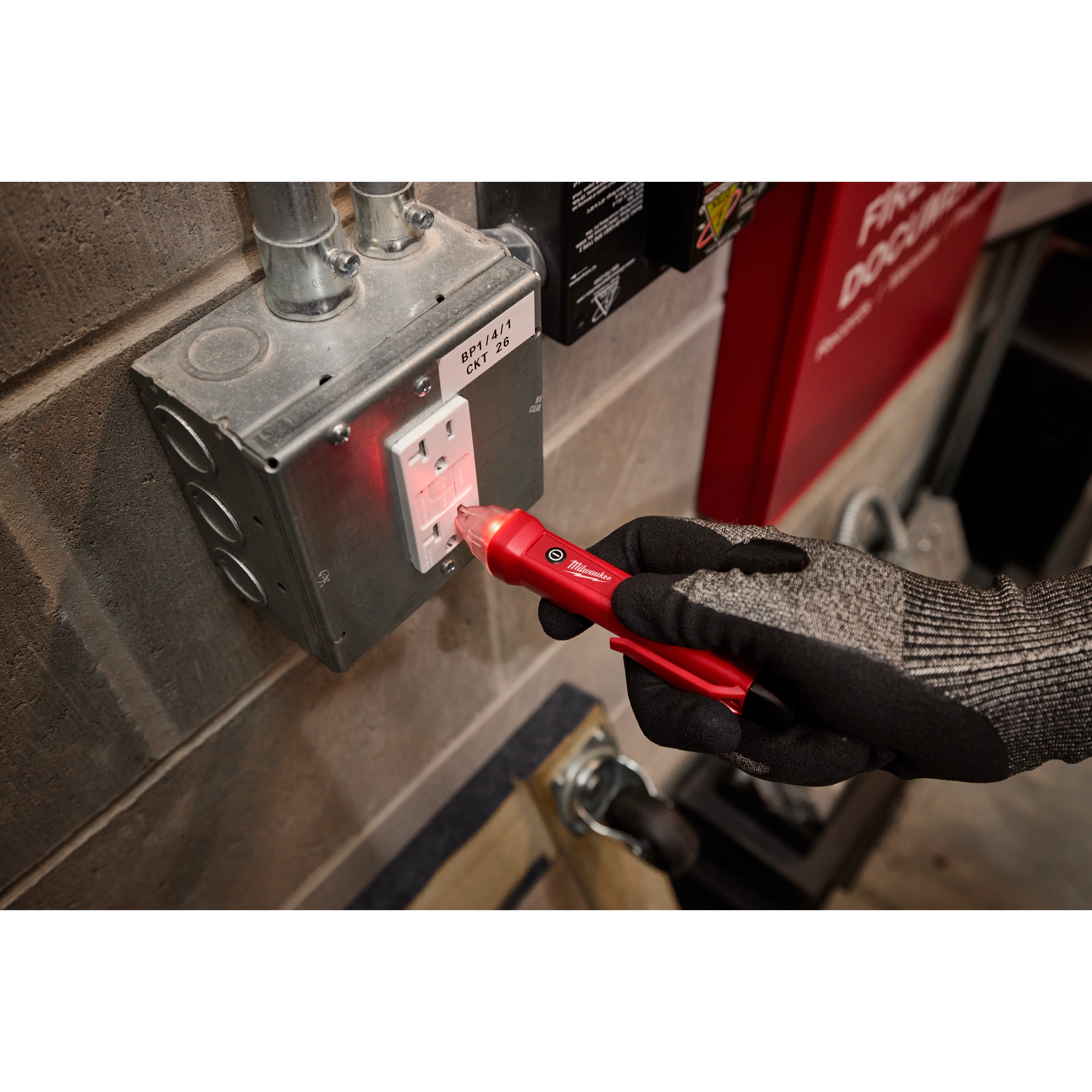 A gloved hand is using a red Voltage Detector to test an electrical outlet in an industrial setting. The outlet is mounted on a metal electrical box with visible conduit pipes above.