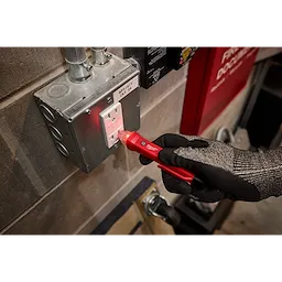 A gloved hand is using a red Voltage Detector to test an electrical outlet in an industrial setting. The outlet is mounted on a metal electrical box with visible conduit pipes above.