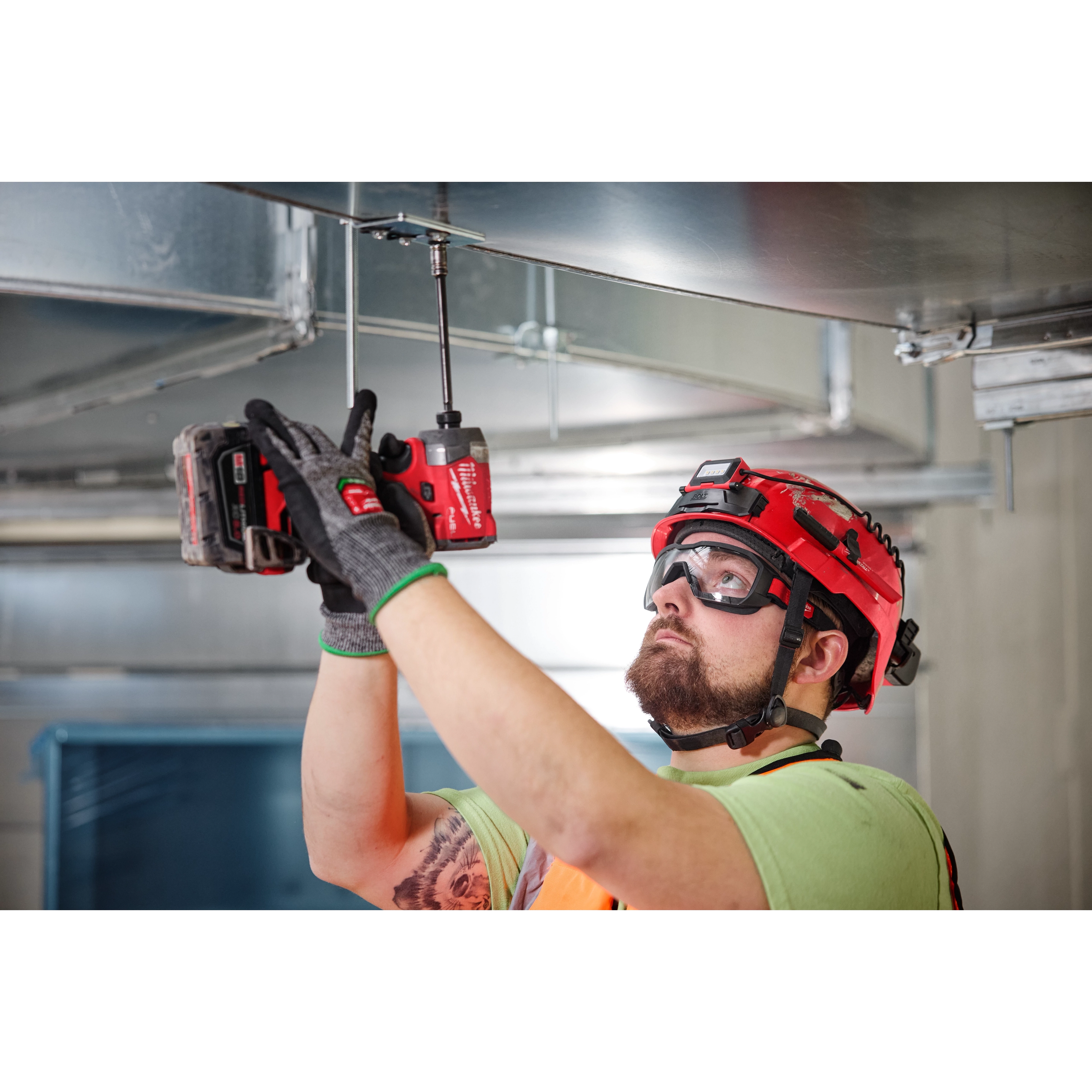 A construction worker wearing Vented Low-Profile Goggles - Clear Dual Coat Lens uses a red cordless drill to work on overhead metal ducting.