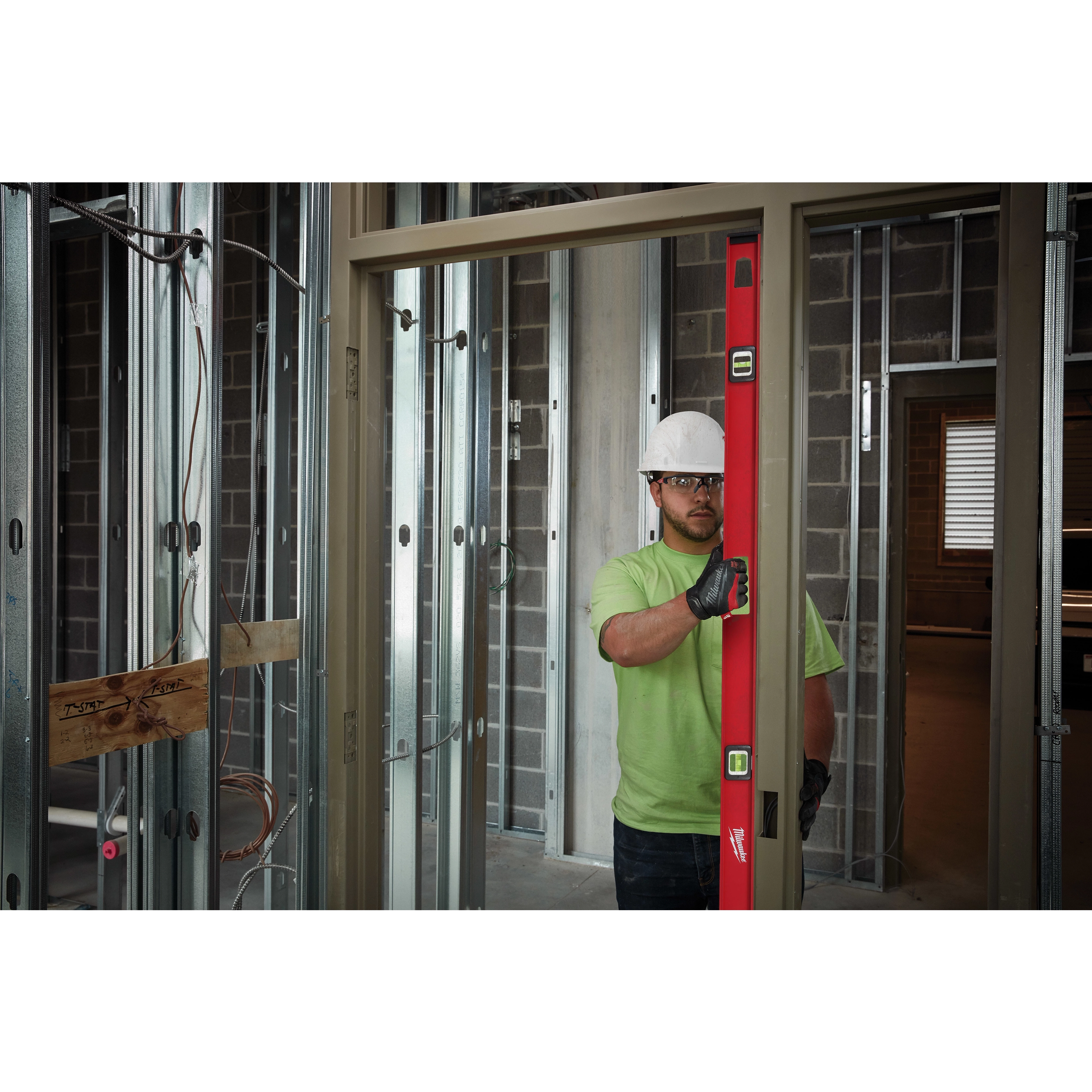 A worker wearing a green shirt and gloves uses a Milwaukee 78" Magnetic I-Beam Level to check the alignment of a door frame in a construction site.