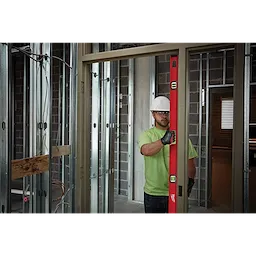 A worker wearing a green shirt and gloves uses a Milwaukee 78" Magnetic I-Beam Level to check the alignment of a door frame in a construction site.