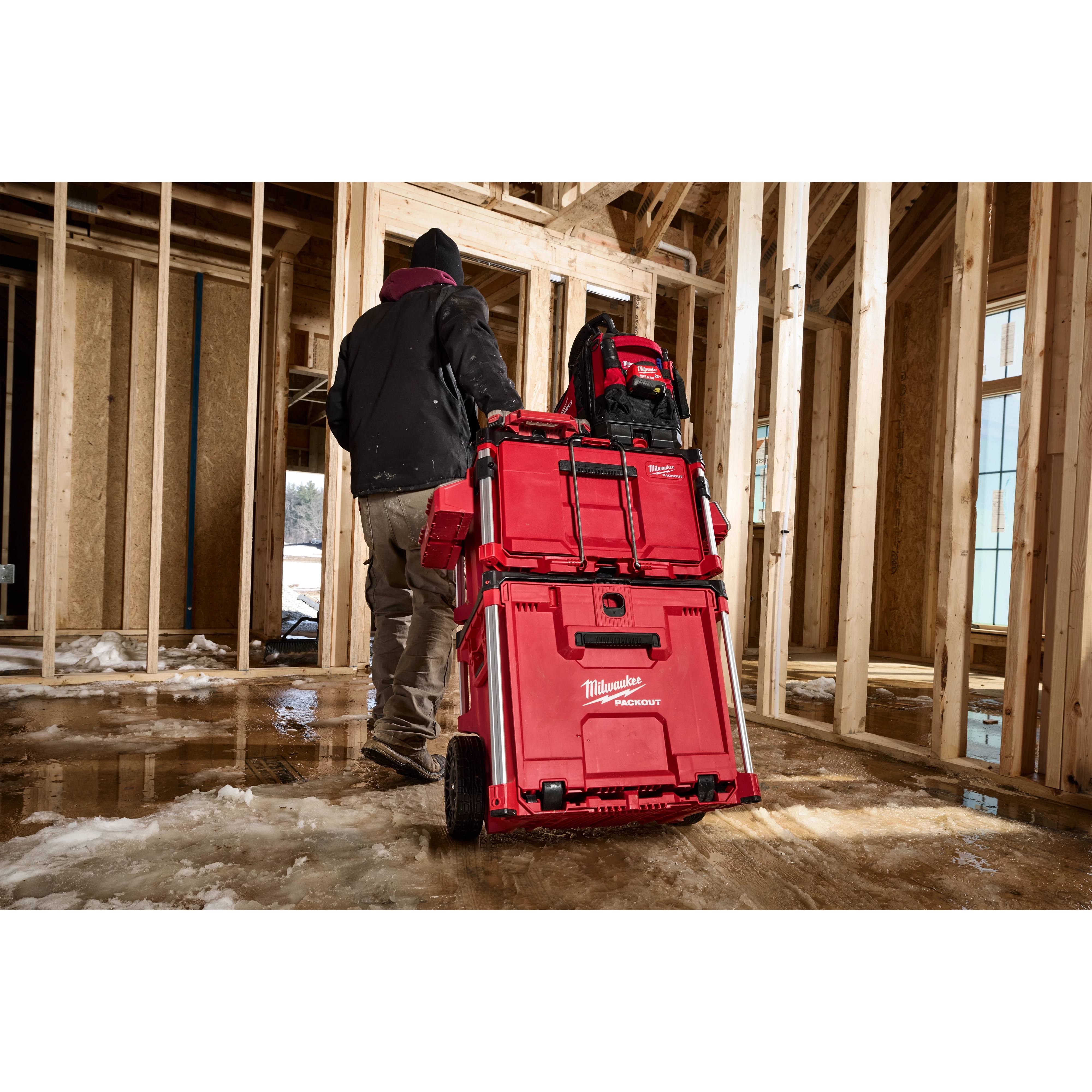 Worker pulling stacked red Milwaukee Packout tool boxes on wheels through a framed building interior.