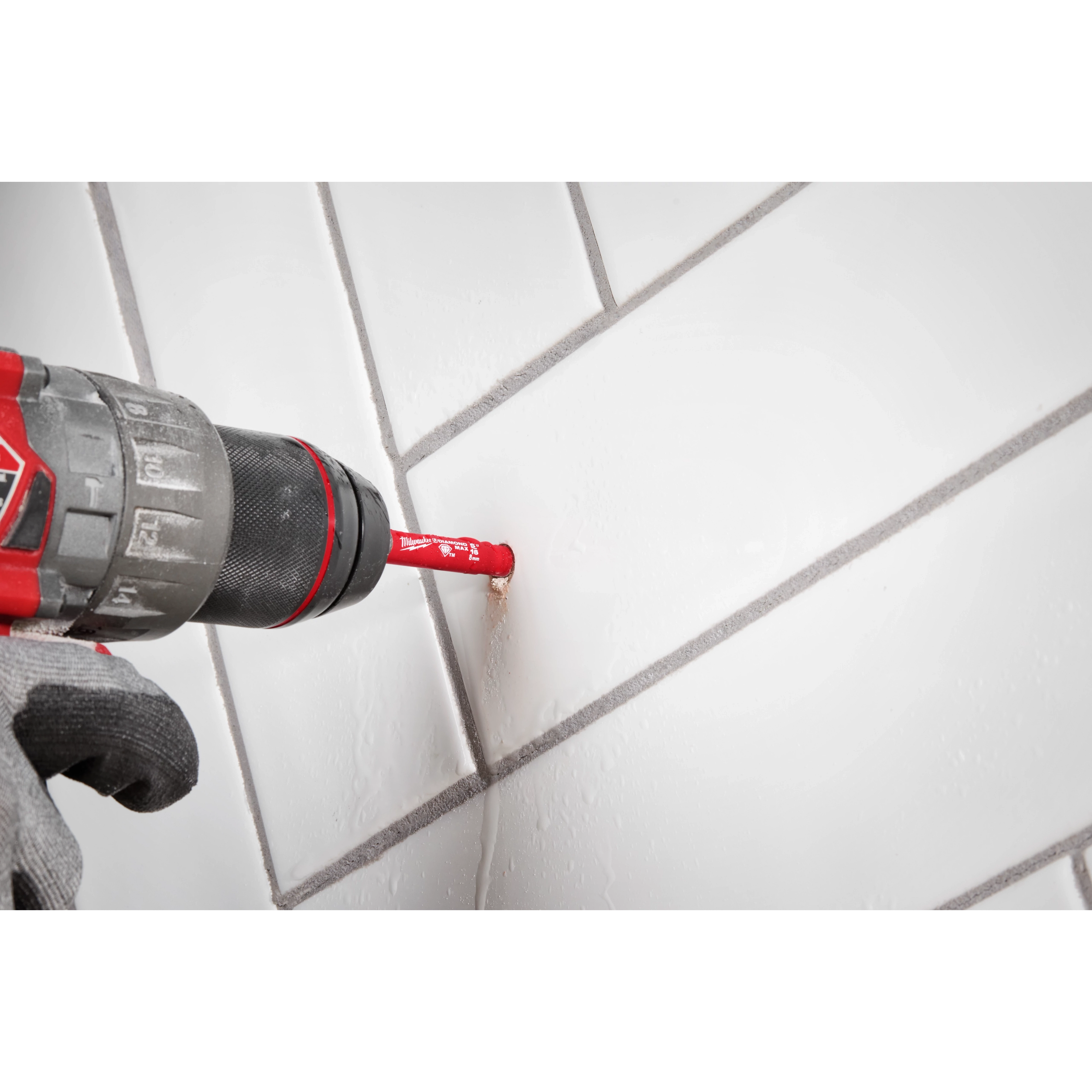 A red Milwaukee® 5/16" DIAMOND MAX™ Hole Saw Bit attached to a black and red power drill is being used to drill a hole in a white tile surface. A gloved hand holds the drill steadily against the tile.