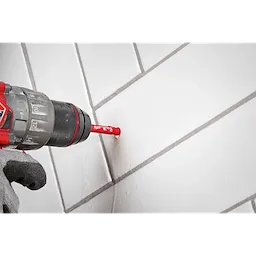A red Milwaukee® 5/16" DIAMOND MAX™ Hole Saw Bit attached to a black and red power drill is being used to drill a hole in a white tile surface. A gloved hand holds the drill steadily against the tile.