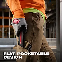 Gloved hand holding Milwaukee Rechargeable Flat Flashlight w/ Green Laser against work pants in an industrial setting.
