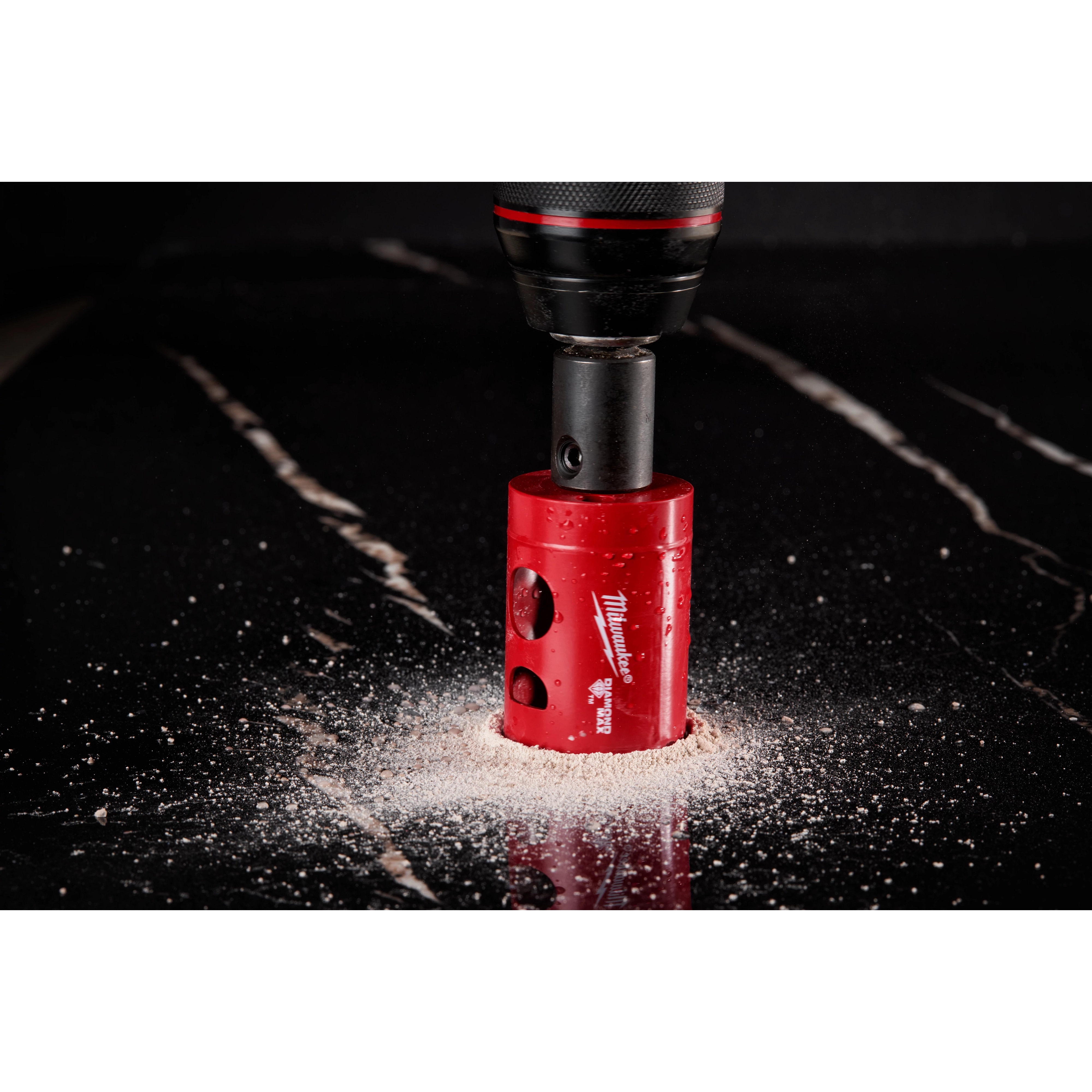 A Milwaukee® 1-3/8" DIAMOND MAX™ Hole Saw is cutting through a dark, polished surface, producing a ring of light-colored dust. The red hole saw is attached to a black drill, showcasing its diamond-enforced teeth designed for durability.