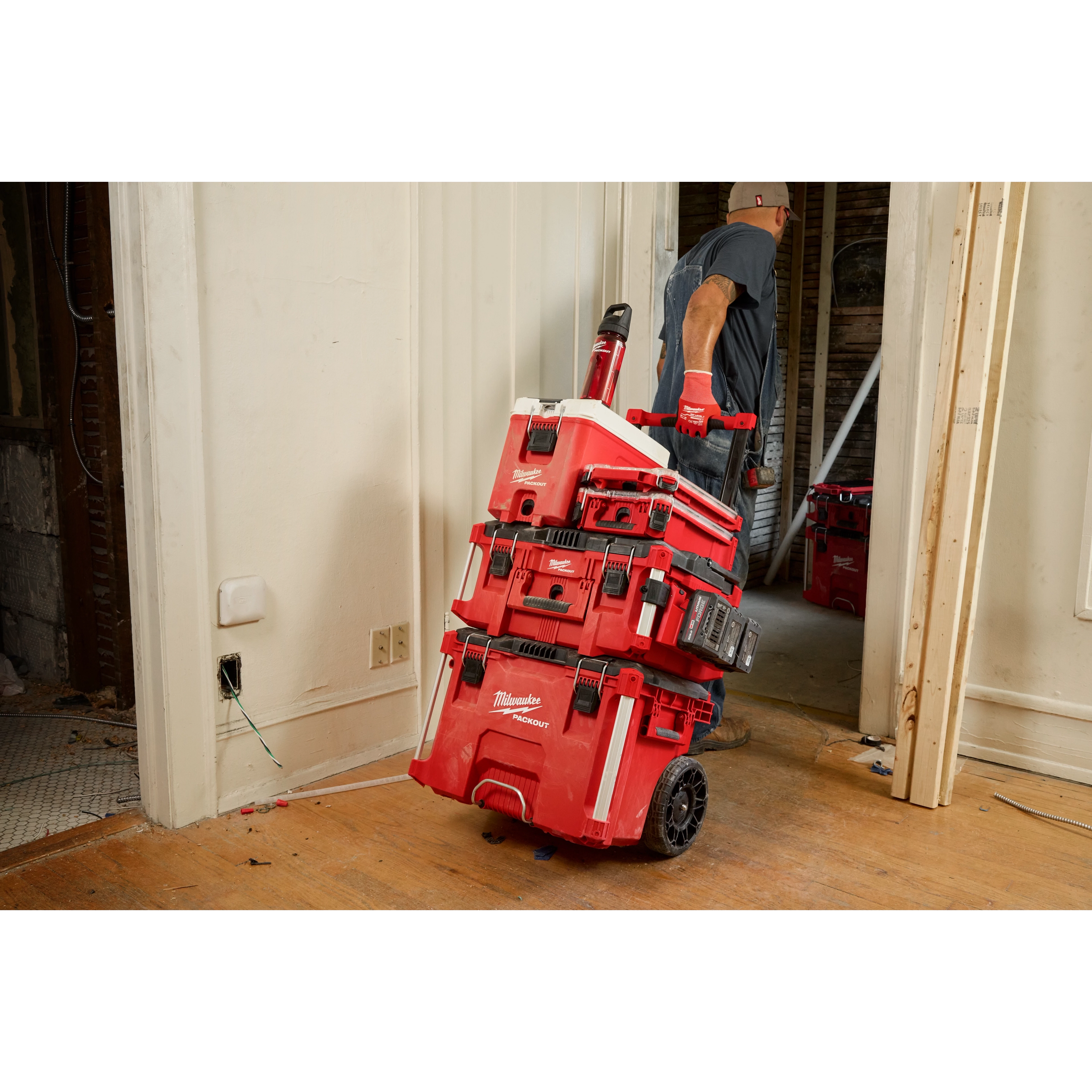 A worker wearing a hard hat and gloves pulls a stacked set of red Milwaukee PACKOUT toolboxes on a wheeled cart through a doorway
