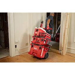 A worker wearing a hard hat and gloves pulls a stacked set of red Milwaukee PACKOUT toolboxes on a wheeled cart through a doorway