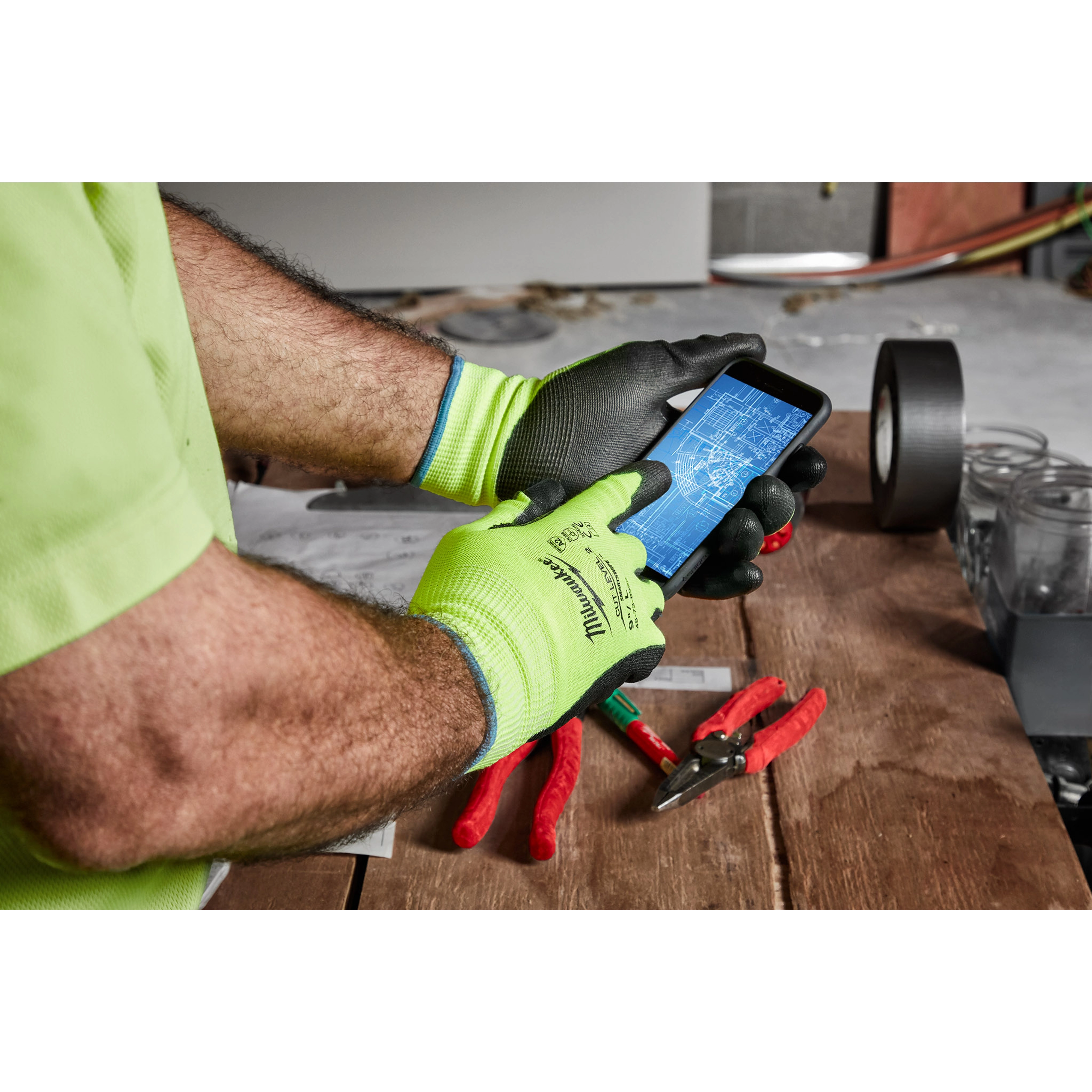 Man wearing Hi-Vis Cut Gloves using a smartphone with pliers and other tools on a wooden table in the background.
