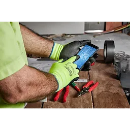 Man wearing Hi-Vis Cut Gloves using a smartphone with pliers and other tools on a wooden table in the background.