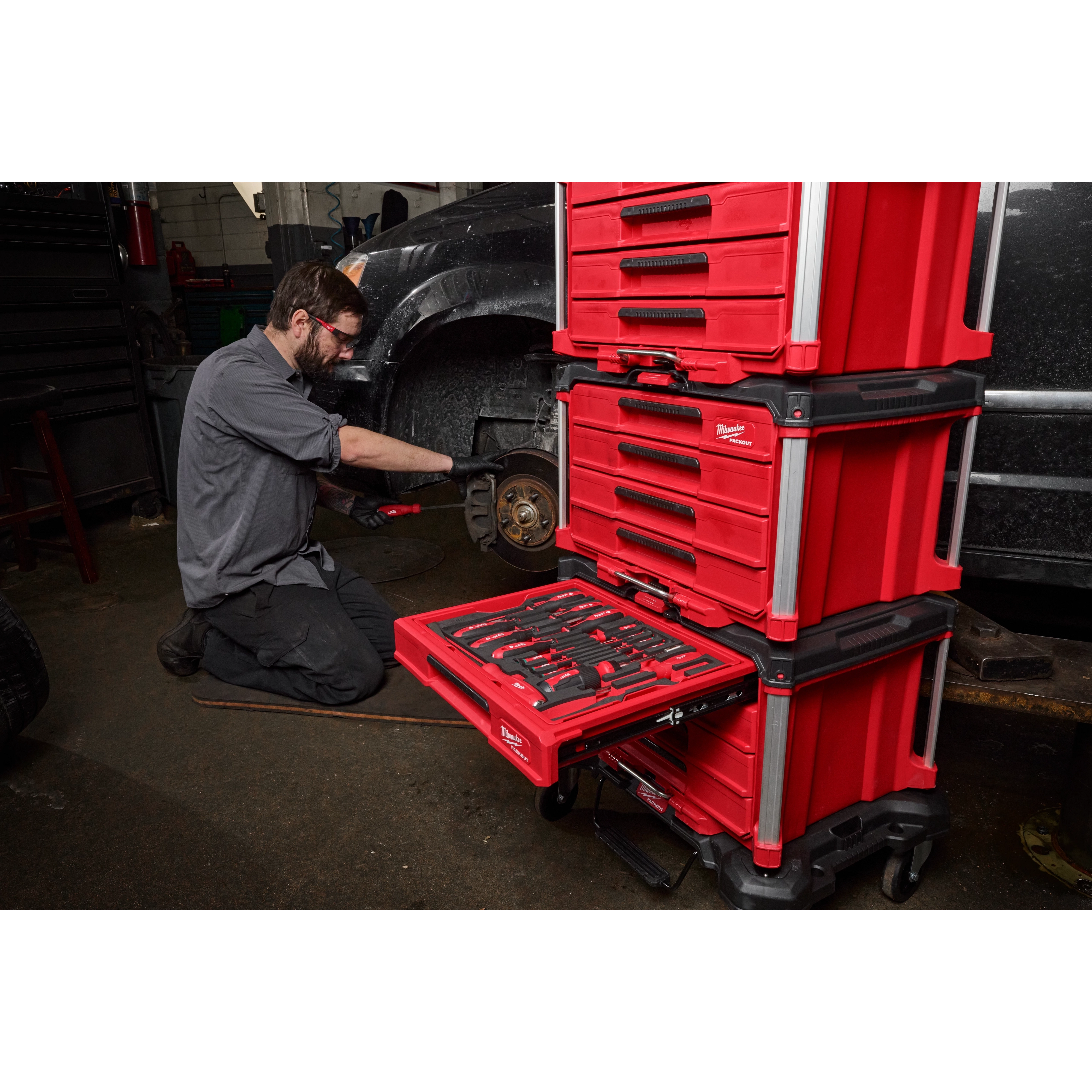 A mechanic utilizes tools from a 366-piece Master Mechanics Hand Tool Set with PACKOUT™ Drawers and Dolly. The red drawer system is stacked and open, showcasing the organized tools while the mechanic works on a vehicle in a garage.