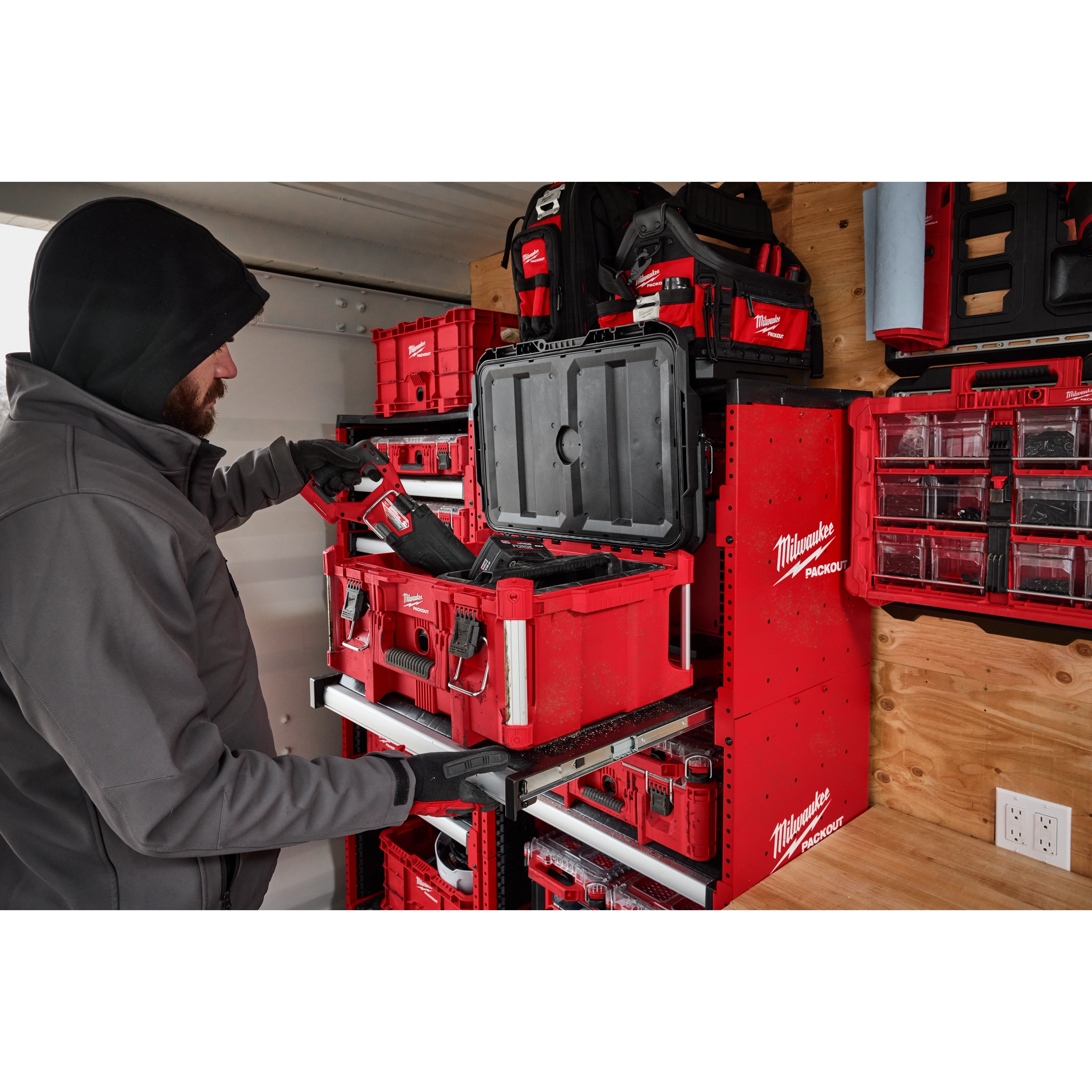 A person uses a PACKOUT™ Rack Kit by Milwaukee, organizing various red and black toolboxes on a mounted shelving unit in a workshop or garage. The rack neatly arranges all the toolboxes and other equipment.