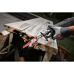 A person uses a 9" 5 TPI AX™ w/NITRUS™ Carbide Teeth SAWZALL® Blade for Wood with Nails to cut through a piece of weathered wood with embedded nails. The blade is attached to a power saw, demonstrating its capability in cutting through tough materials.