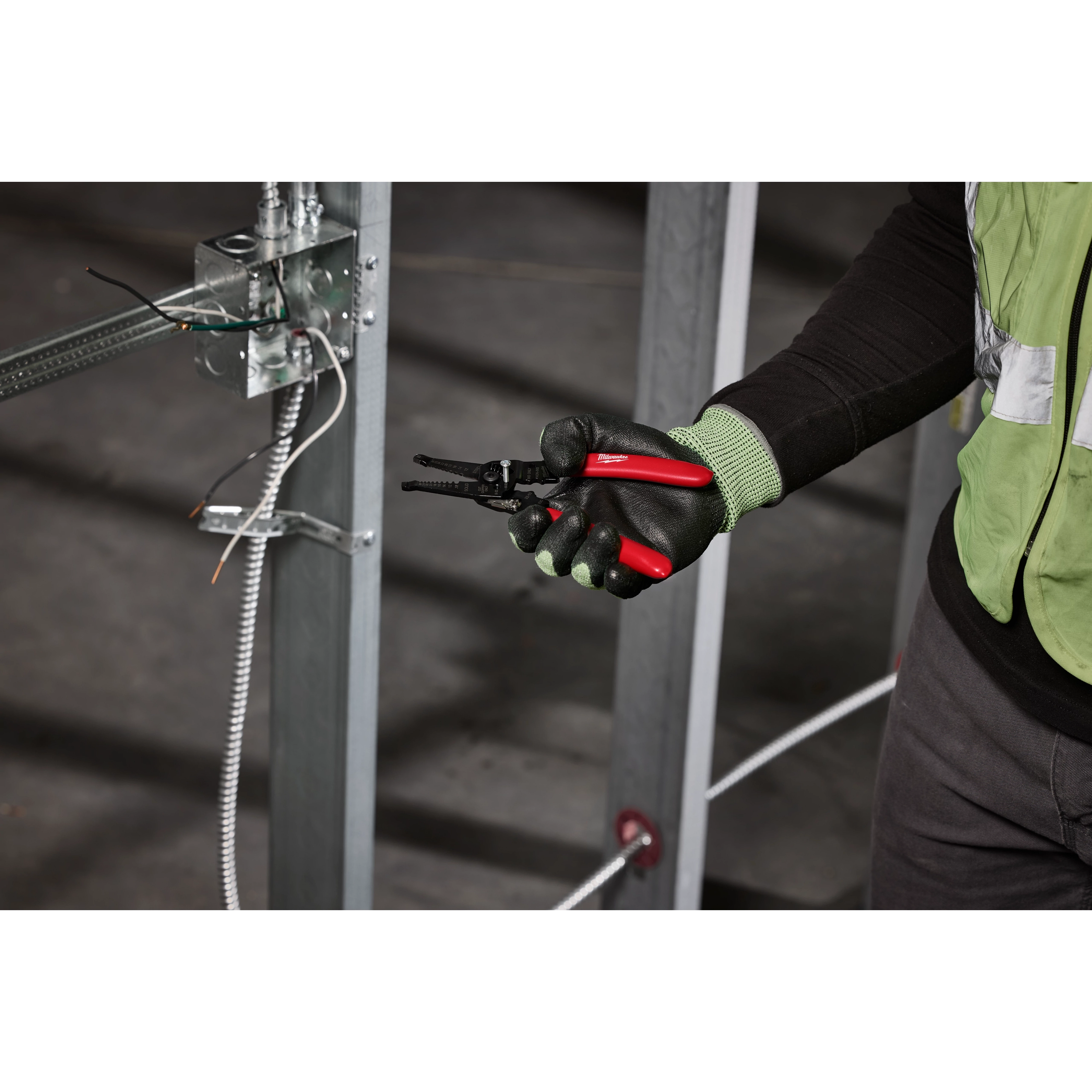 A person wearing gloves holds a 10-28 AWG Multi-Purpose Dipped Grip Wire Stripper & Cutter with Reinforced Head. They are standing near an electrical setup with exposed wires and metal framework.