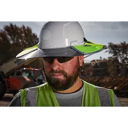 A construction worker wears a white hard hat fitted with a BOLT Sun Visor, featuring bright green accents for increased visibility. The person also wears a grey shirt and a green safety vest.