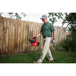 Person walking on grass using an M12 Handheld Broadcast Spreader near a wooden fence.