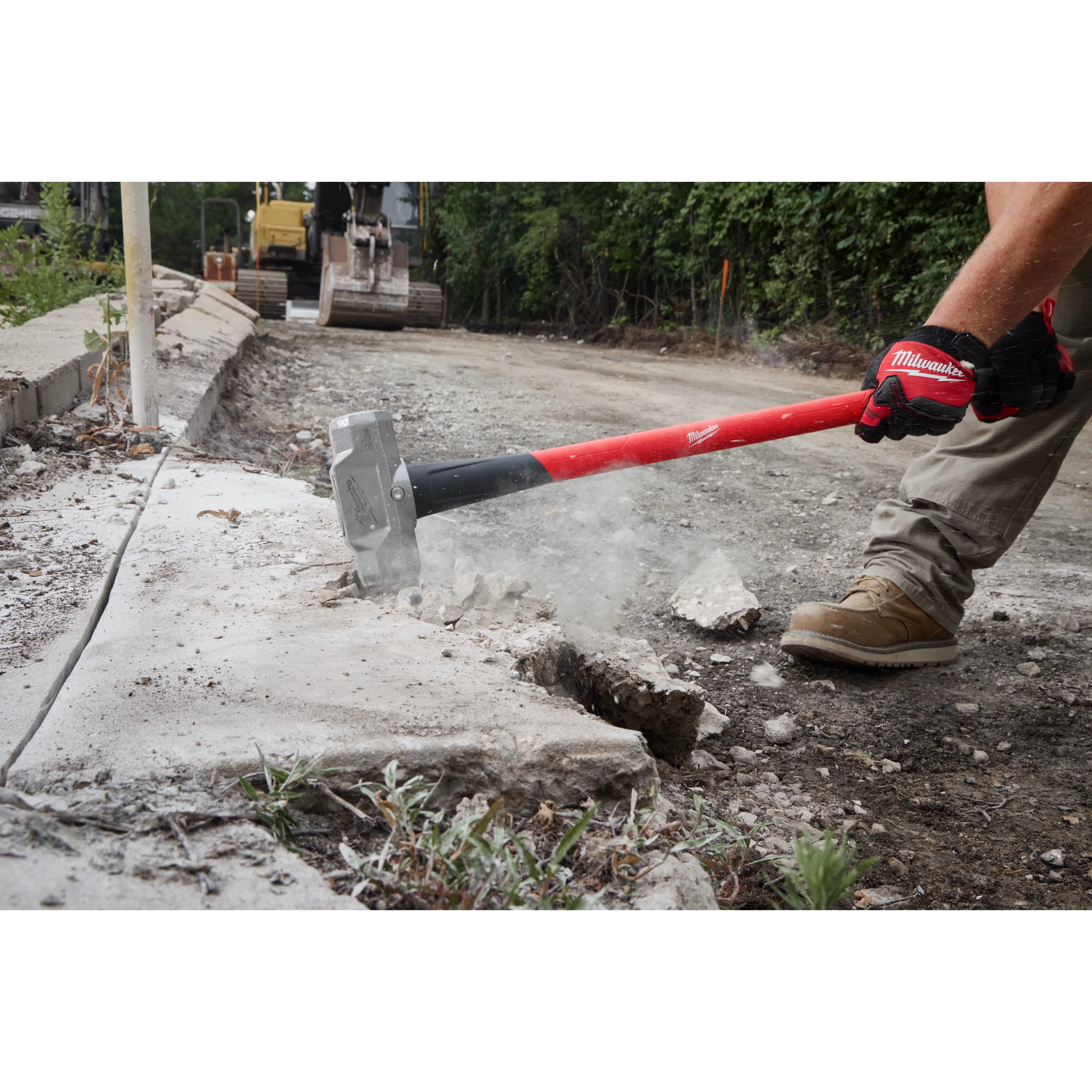 A construction worker uses a 10lb Sledge Hammer (36" Handle) to break apart concrete on a worksite. The sledgehammer has a red handle and its metal head impacts the pavement, causing debris and dust to scatter. The worker wears gloves and sturdy boots, standing on gravel next to heavy machinery.