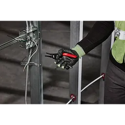 A person wearing gloves holds a 20-32 AWG Low Voltage Dipped Grip Wire Stripper & Cutter. The person is working on an electrical panel with wires.