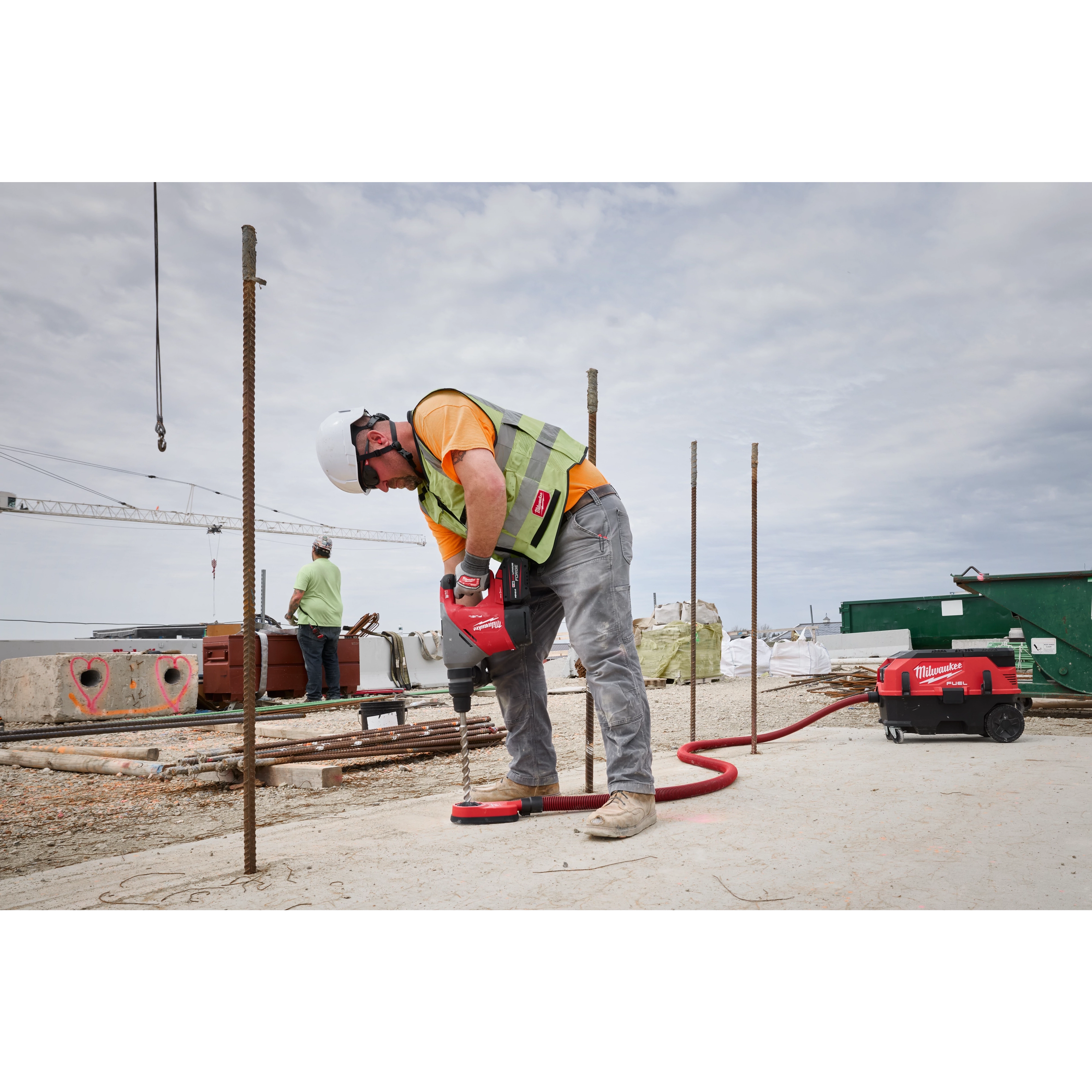 A construction worker uses the M18 FUEL™ 1-9/16" SDS Max Rotary Hammer with ONE-KEY™ to drill into concrete at a job site. The worker wears a safety vest and helmet, and construction materials are visible in the background.