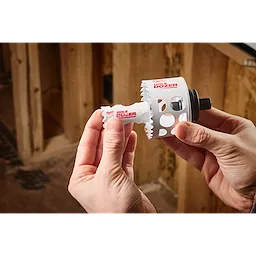 A person holds a Hole Enlargement Adapter for QUIK-LOK™ Arbor tool by Milwaukee in a woodworking shop. The adapter is white with serrated edges and labeled "Hole Dozer."