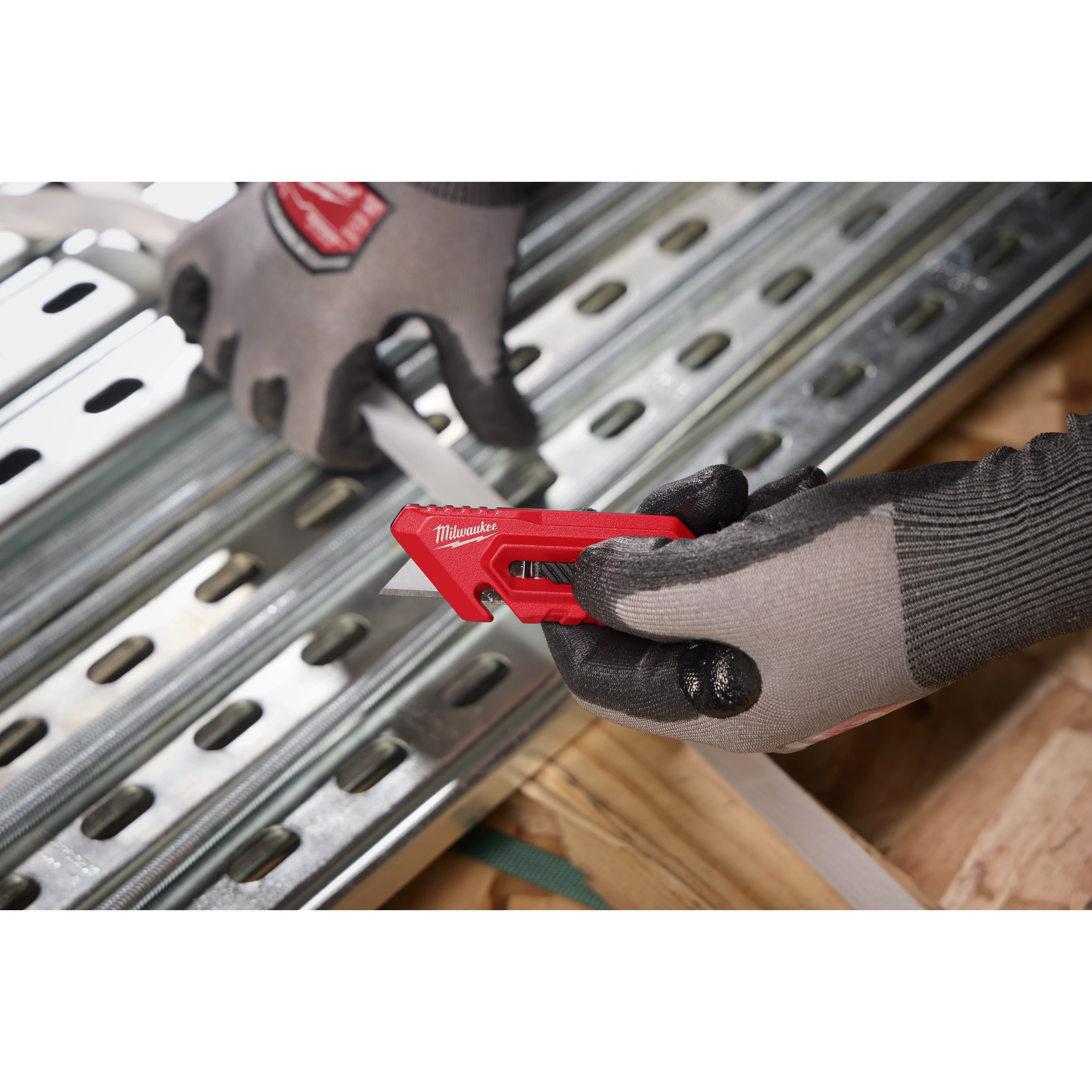 The Multi-function Compact Utility Knife, red with a retractable blade, is held by a gloved hand. Another gloved hand holds a white strip against a metal sheet. The knife is used for precise cutting in construction or DIY projects.