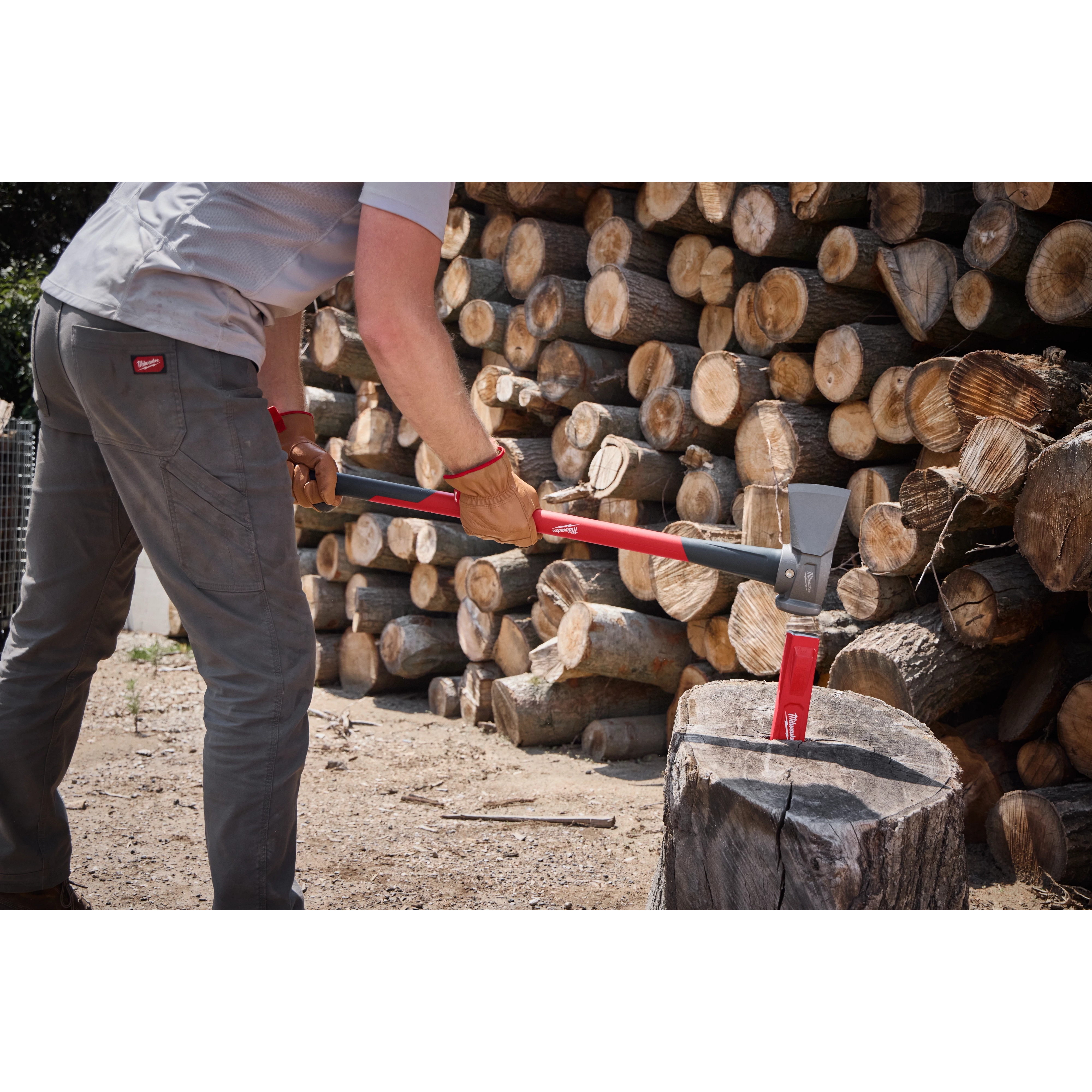 A person uses an 8lb Splitting Maul (36" Handle) to split a large piece of wood. The maul features a grey and red handle and is shown in interaction with a log. There are stacks of cut logs in the background. The person is wearing grey pants and a light shirt.