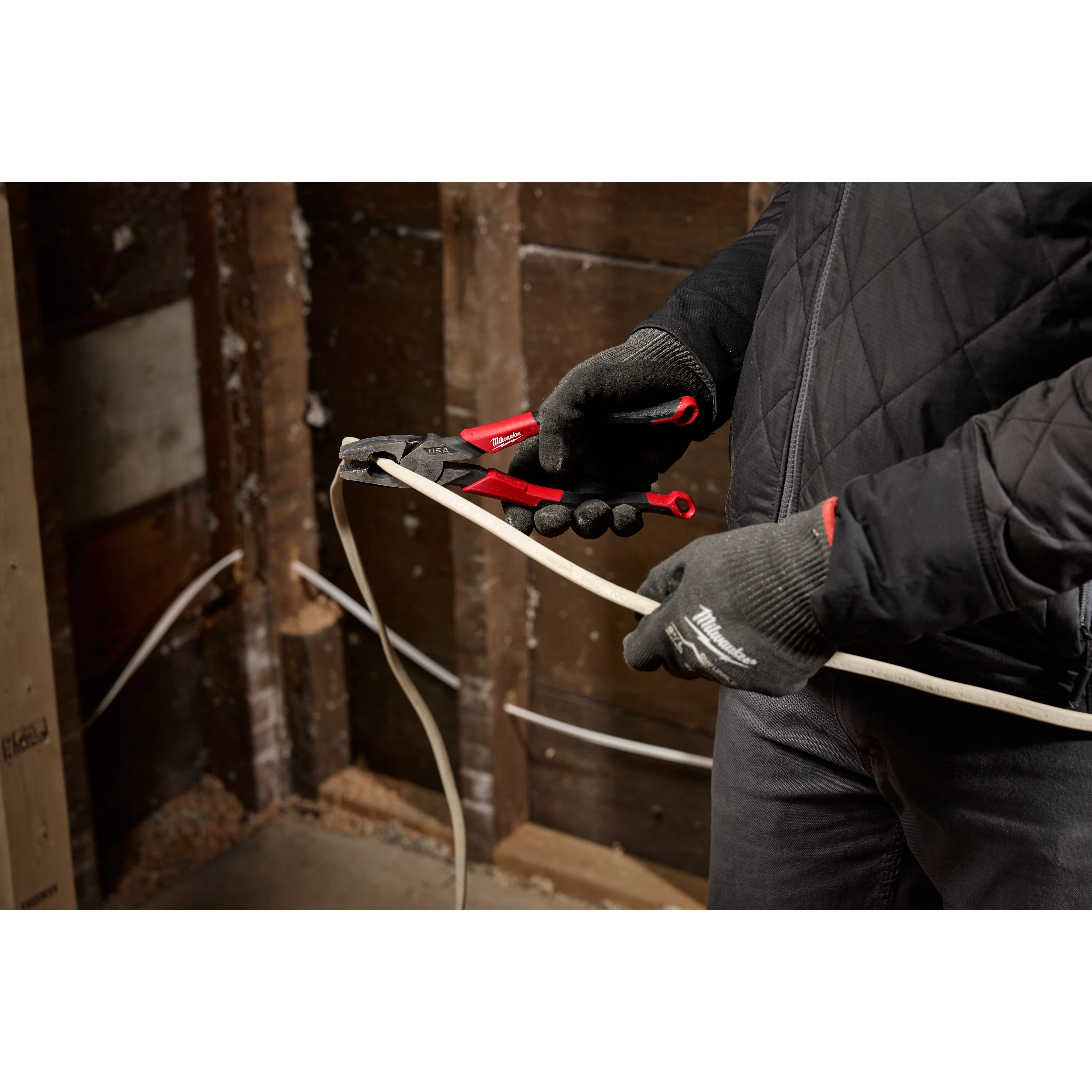 A person wearing gloves and a black jacket uses a pair of red-handled pliers to crimp an electrical wire inside a wooden stud wall under construction.