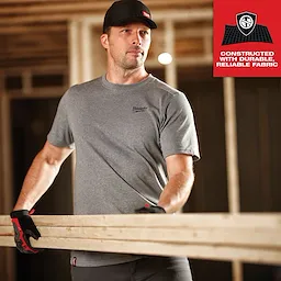 Man wearing GRIDIRON Snapback Trucker Hat - Black, gray t-shirt, and work gloves, holding a wooden plank at a construction site.