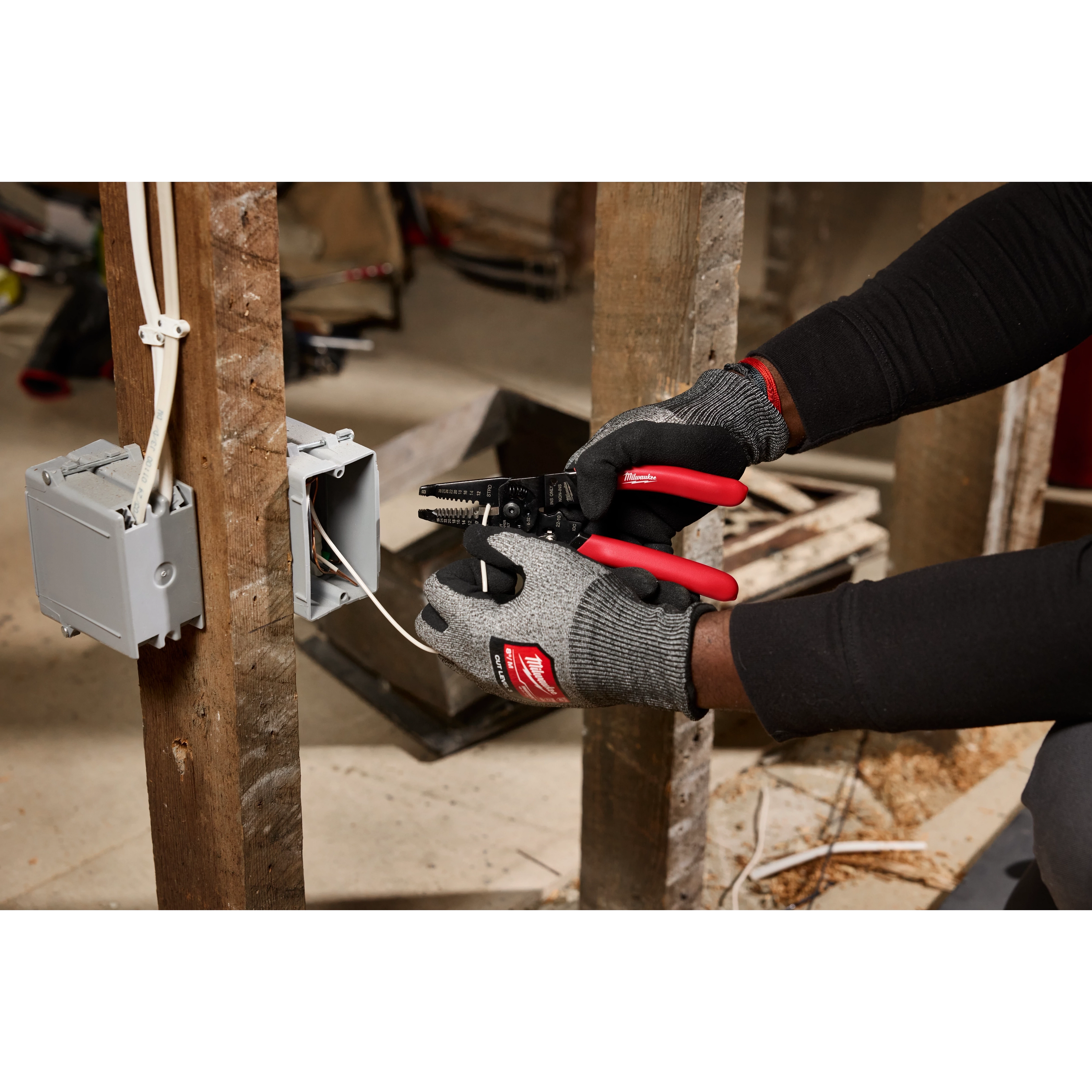 A person wearing gloves uses a 10-28 AWG Multi-Purpose Dipped Grip Wire Stripper & Cutter with Reinforced Head to strip wires in an electrical outlet box on a wooden stud.