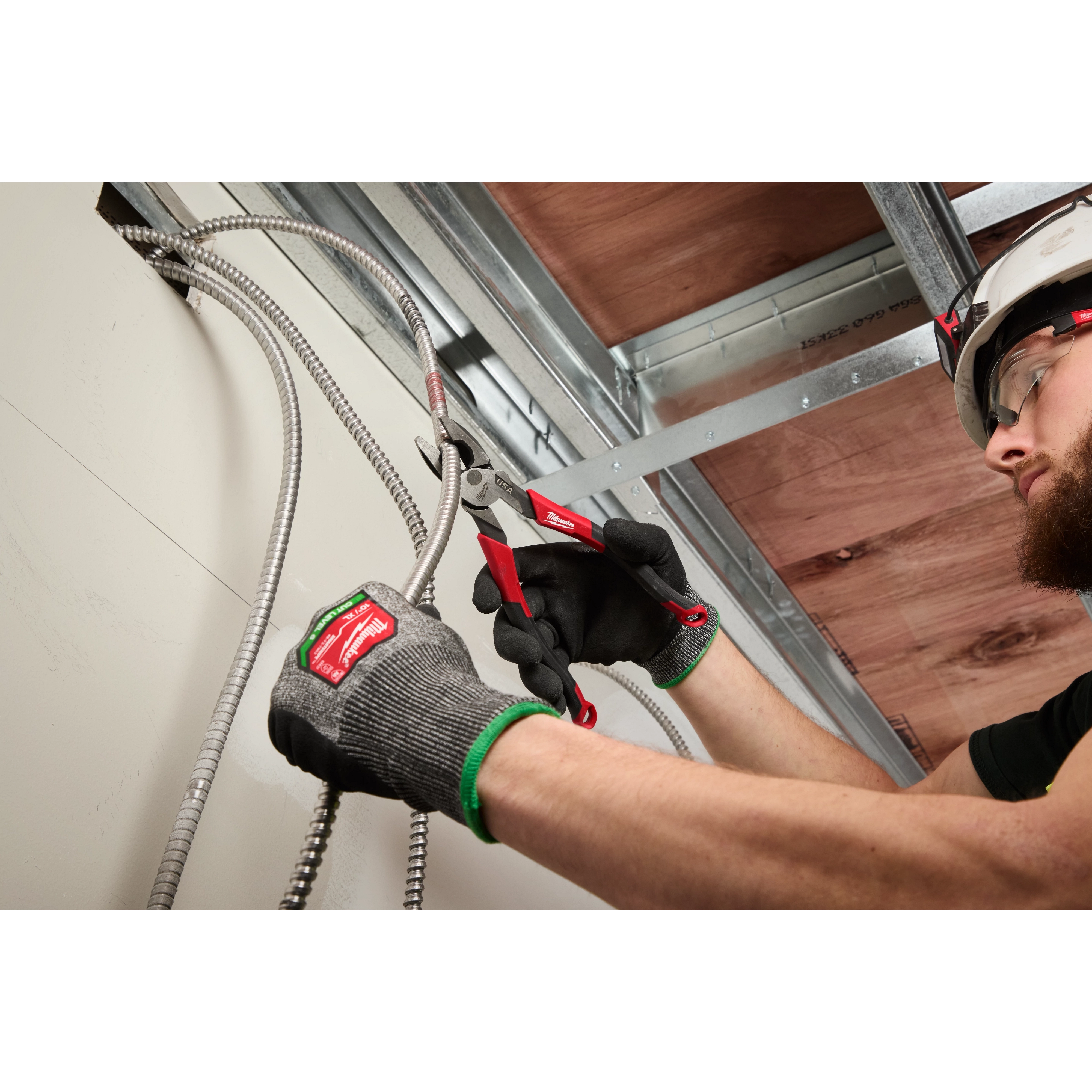 A person wearing green and black gloves uses red-handled pliers to cut flexible metal conduit wires in a space with exposed ceiling beams. The person is working precisely and appears focused on the task.