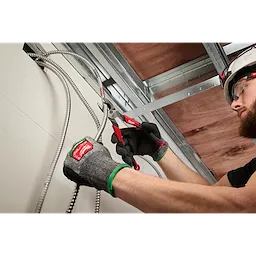 A person wearing green and black gloves uses red-handled pliers to cut flexible metal conduit wires in a space with exposed ceiling beams. The person is working precisely and appears focused on the task.