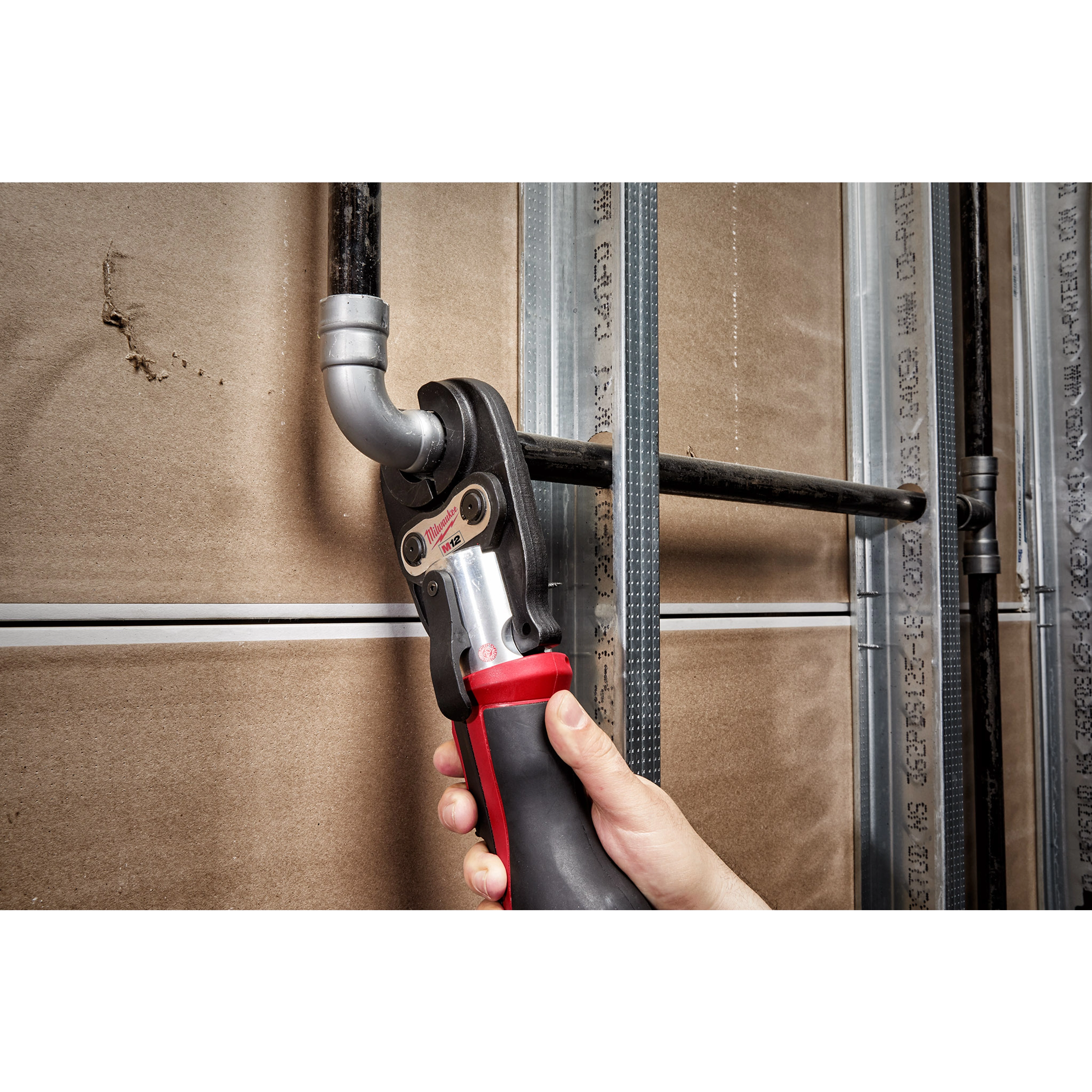 A person uses a 3/4" IPS Jaw for M12 FORCE LOGIC Press Tool to secure a metal pipe fitting against a drywall backdrop.