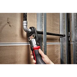 A person uses a 3/4" IPS Jaw for M12 FORCE LOGIC Press Tool to secure a metal pipe fitting against a drywall backdrop.