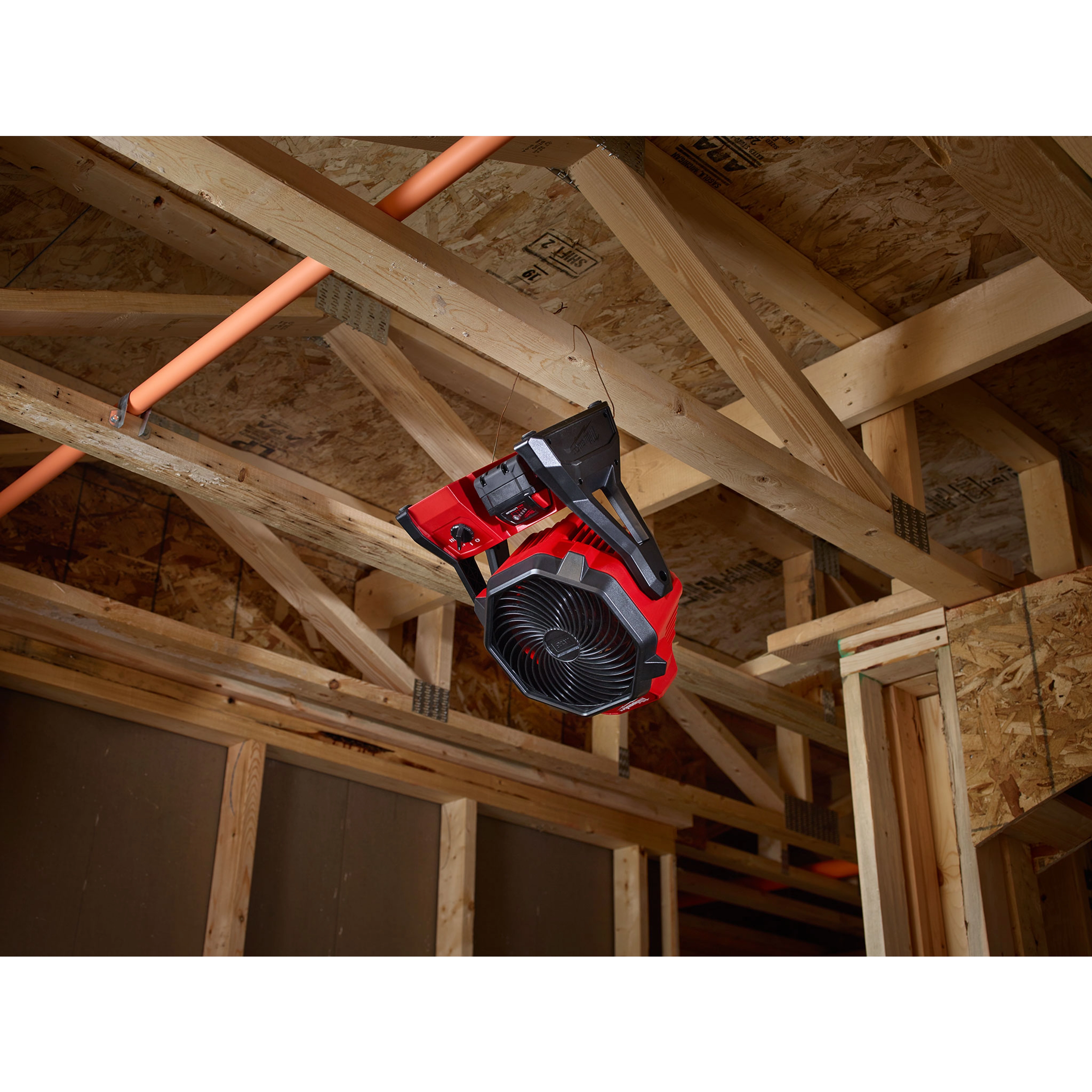 Red and black M18 Jobsite Fan mounted on the wooden frame of a ceiling in a construction site.