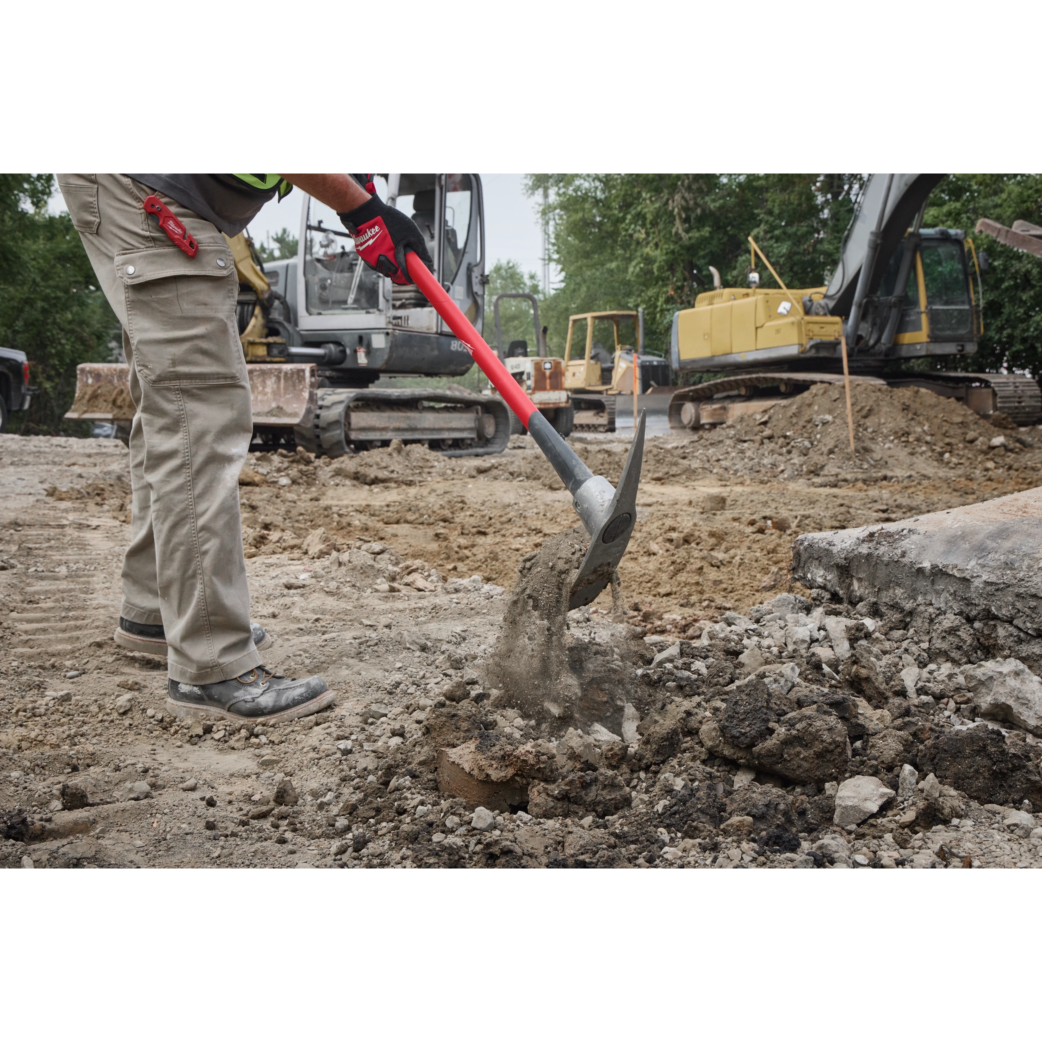 A person uses a 5lb Pick Mattock with a 36" handle to break up hard ground at a construction site. Heavy machinery and earth movers are visible in the background. The user wears protective gloves and work pants.