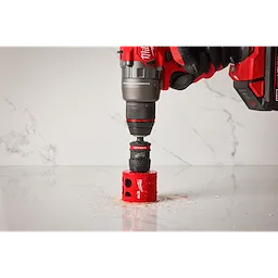 A red and black Milwaukee electric drill with a 2PK Large Thread Pin Adapter for QUIK-LOK™ Arbor attached is drilling into a white marble surface. The drill bit is surrounded by red shavings.