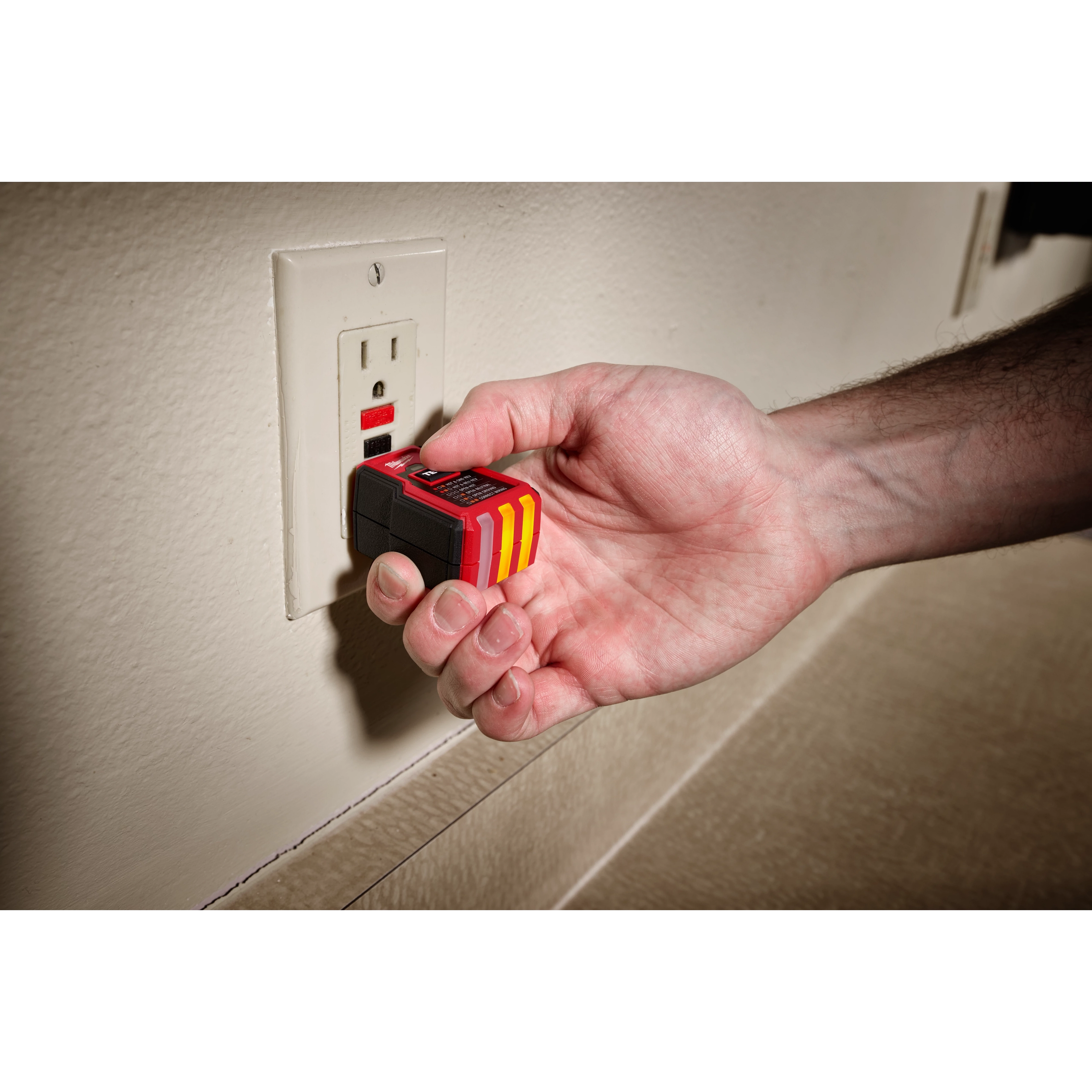A person's hand is holding a GFCI Receptacle Tester, plugging it into a wall outlet. The tester device is black and red with yellow stripes. The outlet has a built-in GFCI reset button, indicating it provides ground fault circuit interrupter protection.