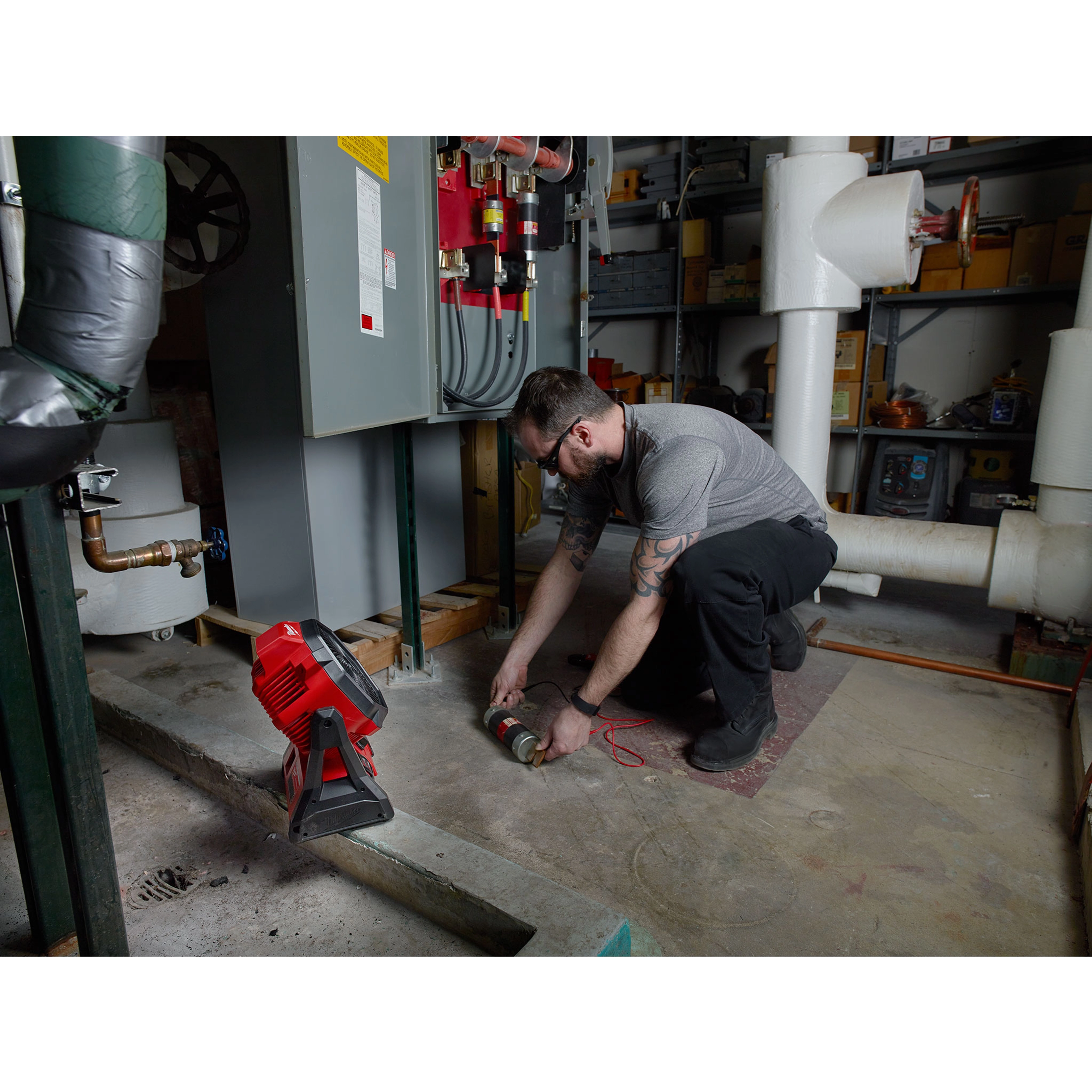 Man using a wrench near machinery in an industrial setting with an M18 Jobsite Fan placed on the floor nearby.