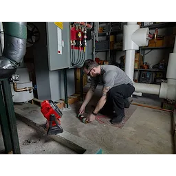 Man using a wrench near machinery in an industrial setting with an M18 Jobsite Fan placed on the floor nearby.