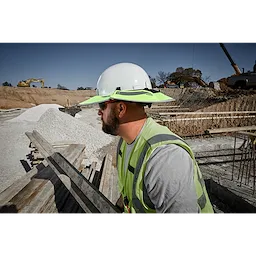 A construction worker in a safety vest and hard hat equipped with the BOLT Sun Visor is at a worksite with earth-moving equipment and materials.
