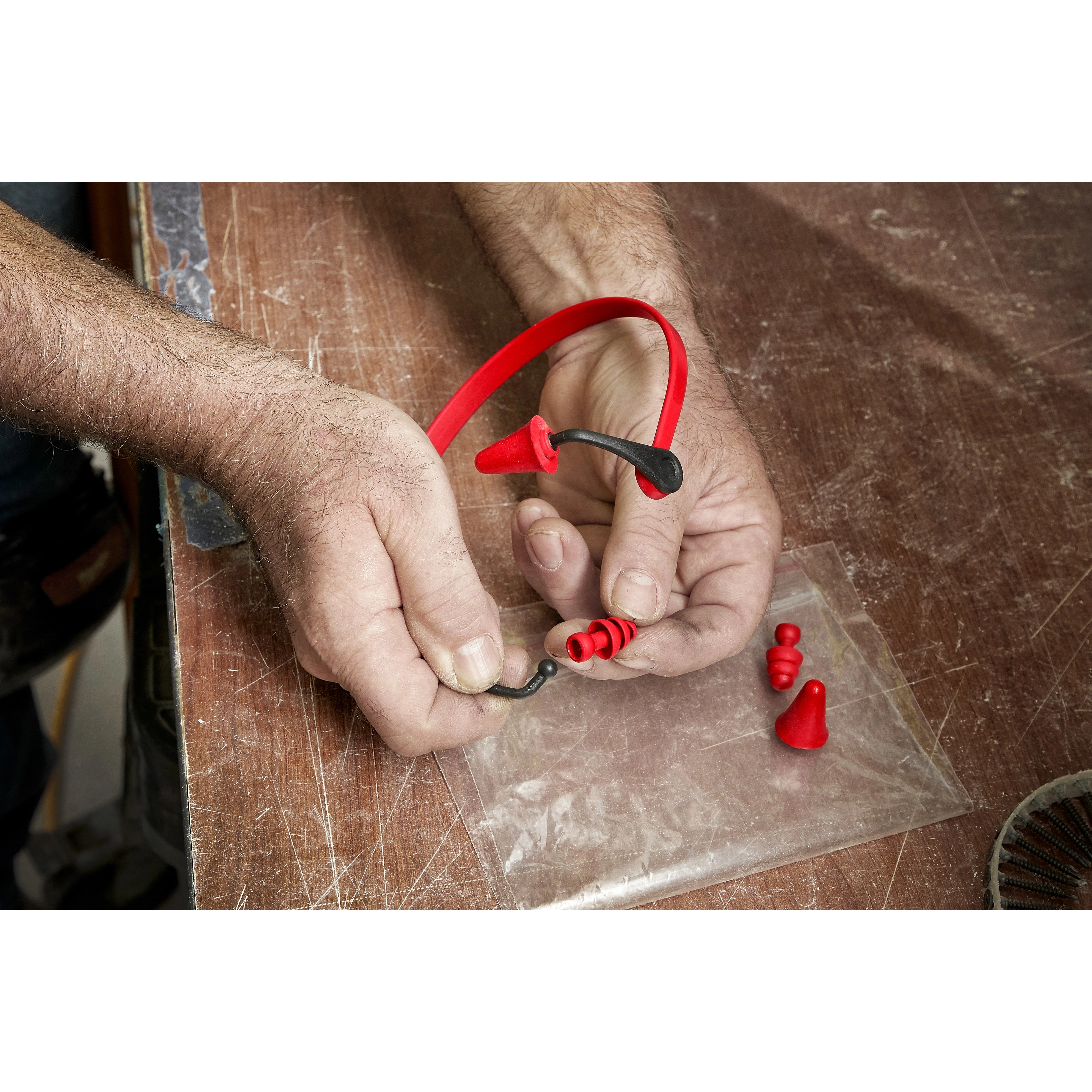 A pair of hands assembling Banded Hearing Protection, a red and black band with replaceable earplugs, on a wooden surface.
