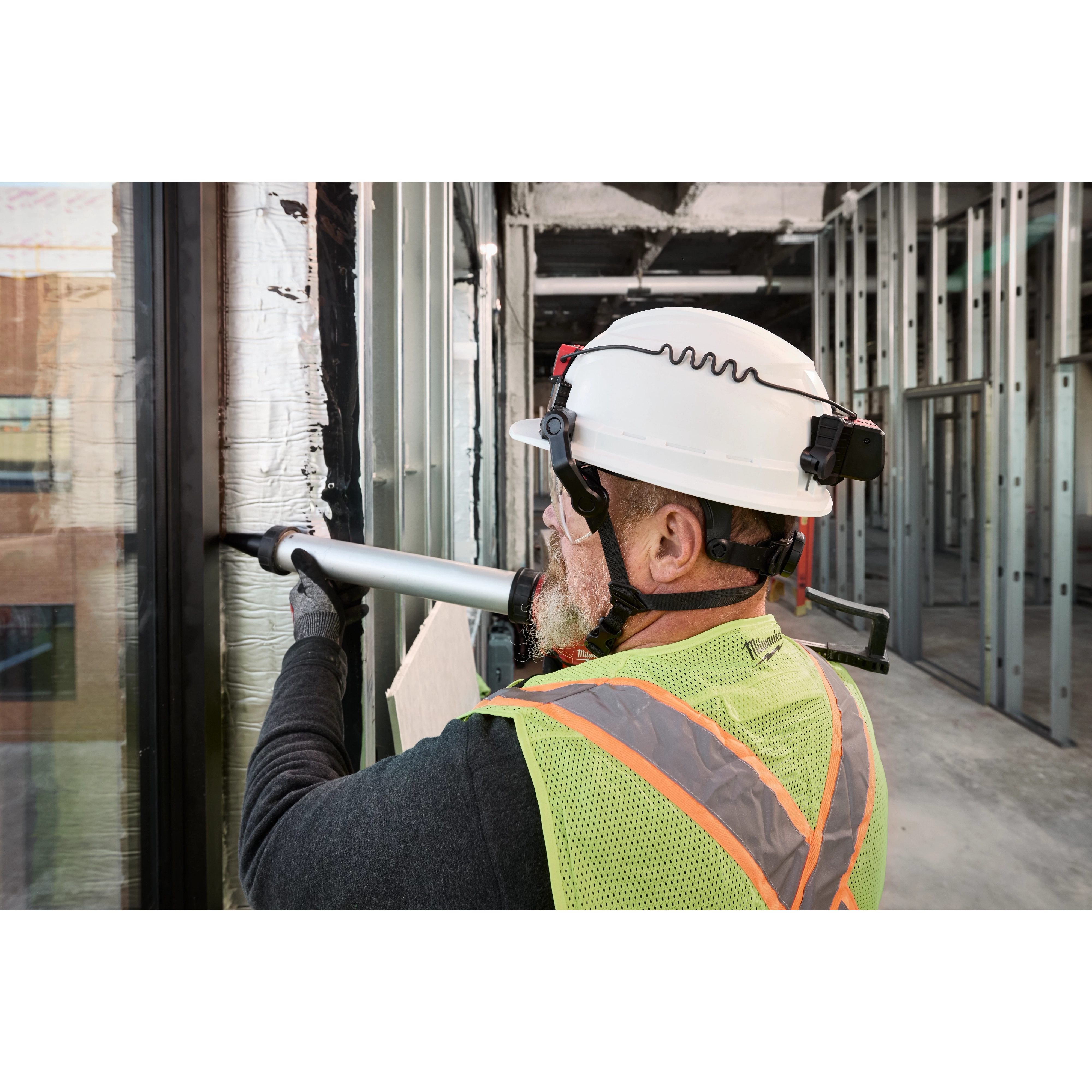A construction worker wearing a BOLT 4PT White Front Brim Non-Vented Safety Helmet, a hi-vis vest, and using a power tool on a building frame.