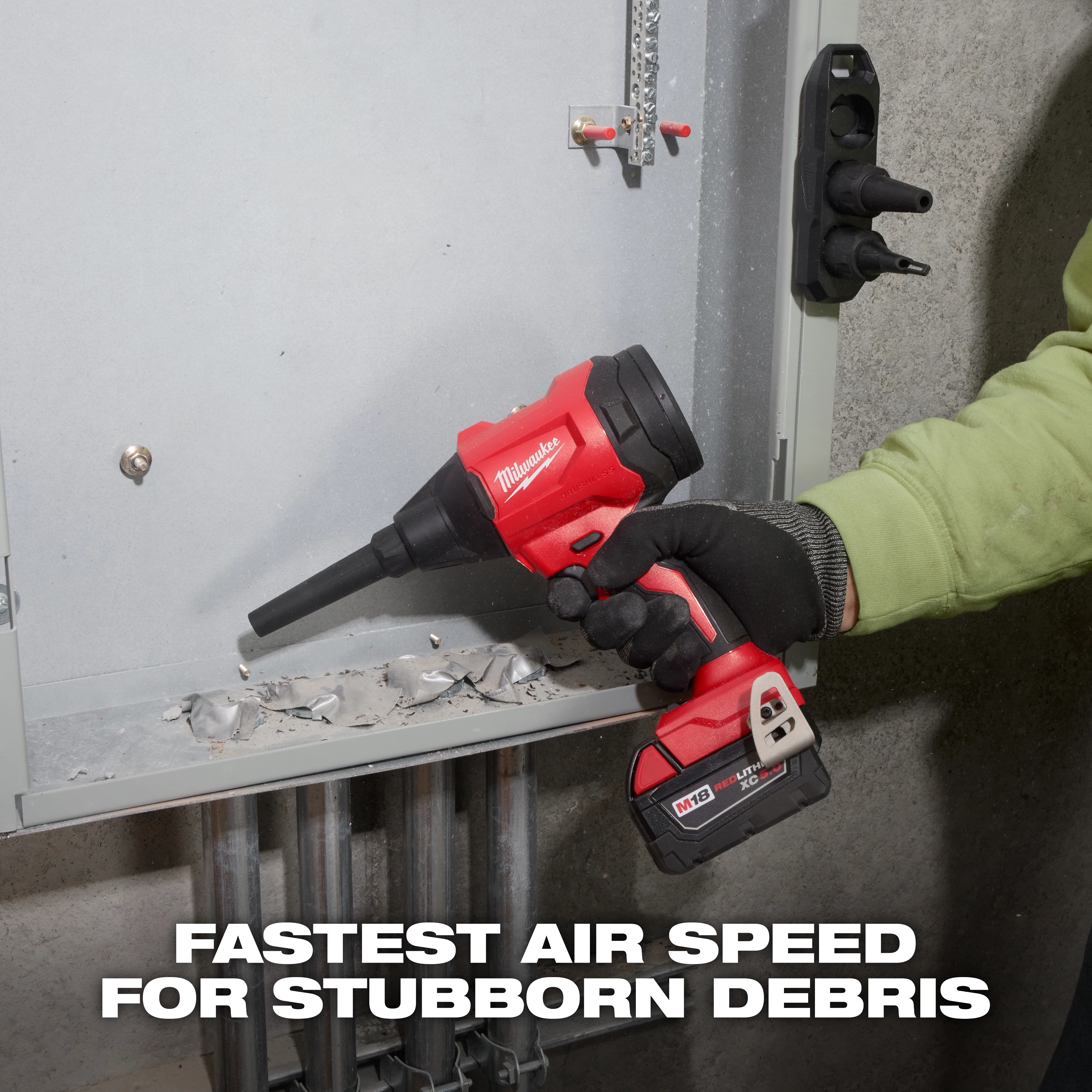 A person uses the M18™ Brushless Precision Blower in a maintenance setting. They are wearing gloves and clearing debris from a metal surface inside an electrical panel. The blower is red and black with a nozzle attachment. Text reads "FASTEST AIR SPEED FOR STUBBORN DEBRIS."