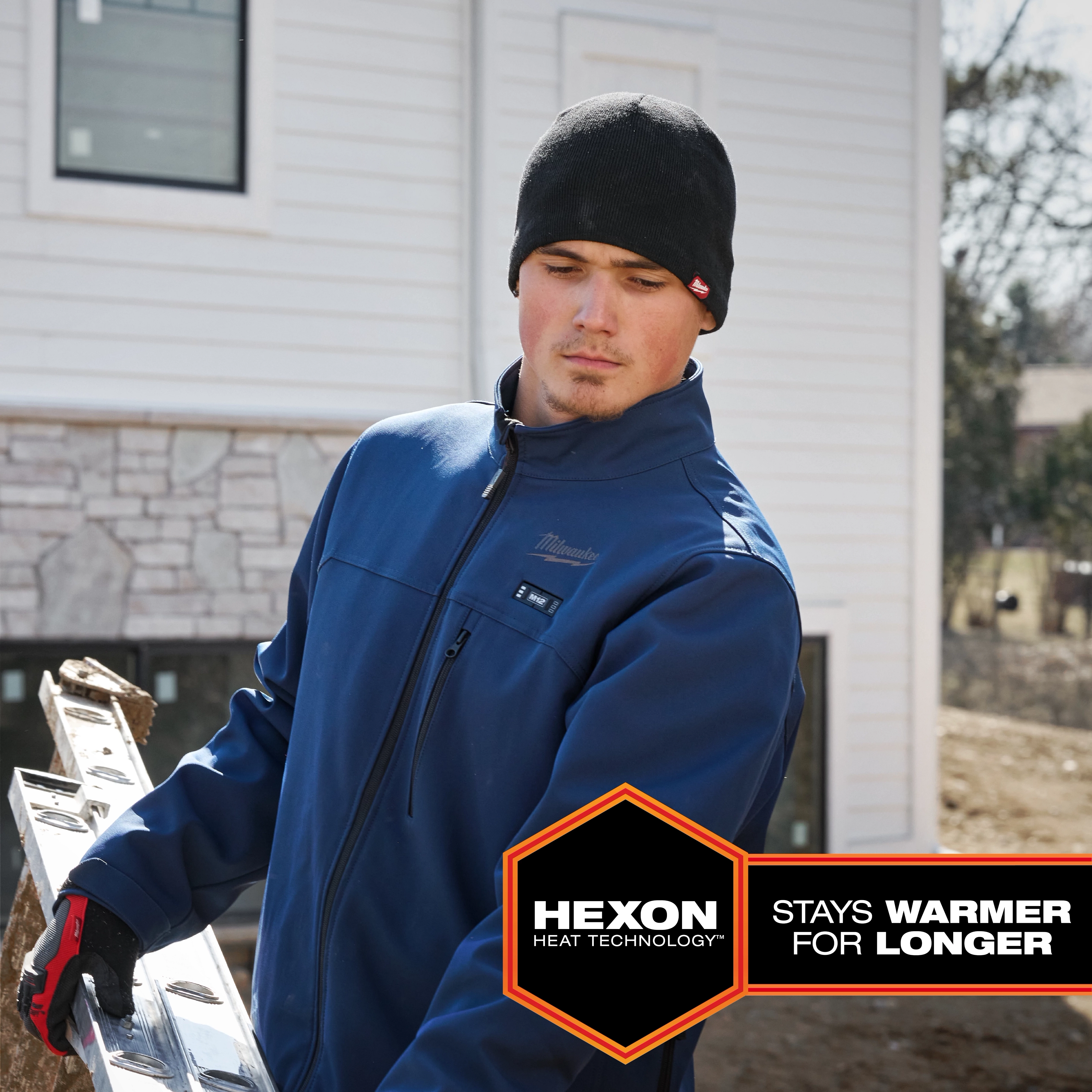 A person wears a blue M12 Heated TOUGHSHELL Jacket while carrying a ladder outdoors. The text reads "HEXON Heat Technology: Stays Warmer for Longer."