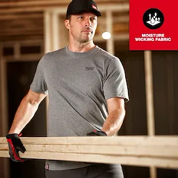Man wearing a grey FREEFLEX Hybrid Work Tee - Short Sleeve while carrying a wooden plank, with moisture-wicking fabric text overlay.