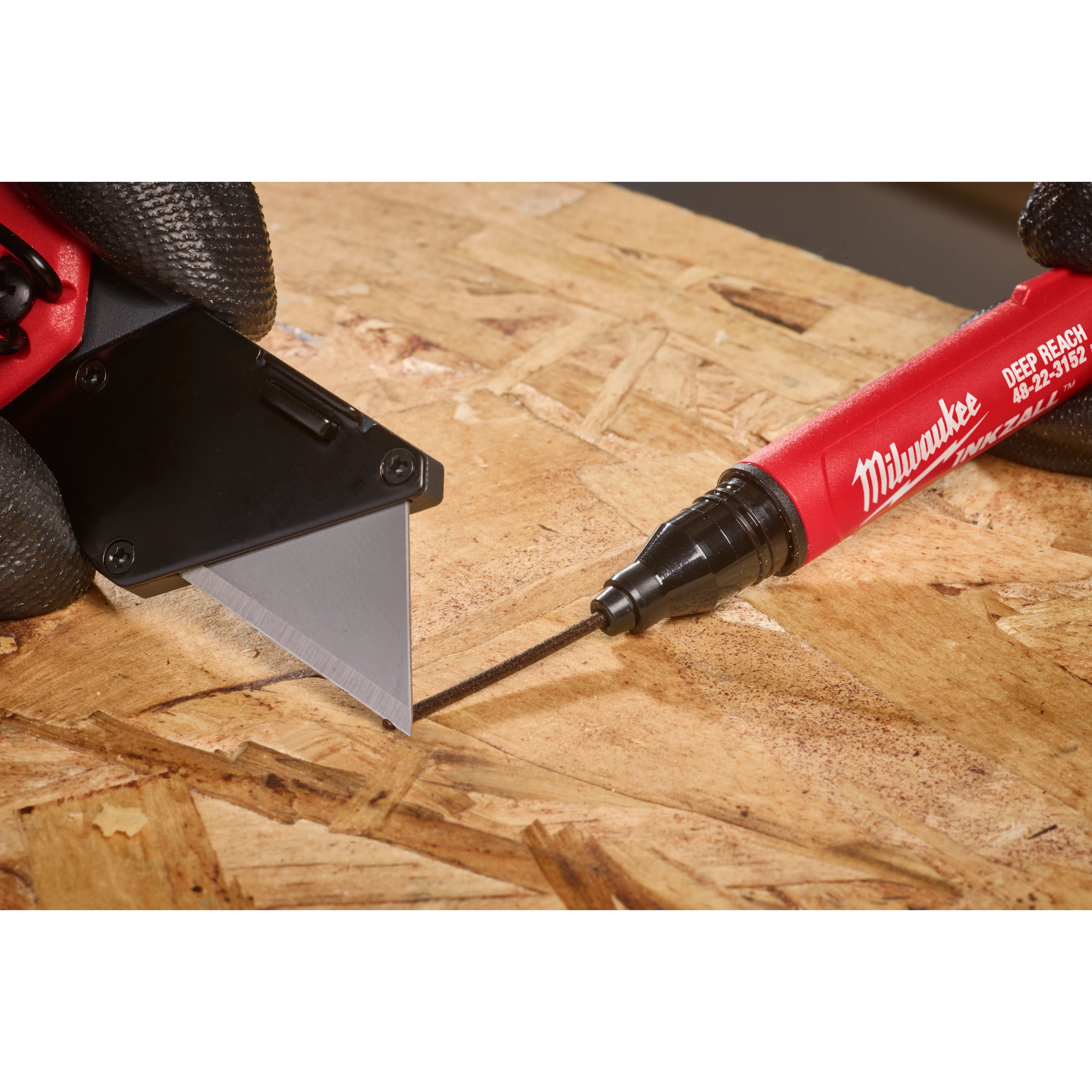 The INKZALL™ 2IN1 Deep Reach Ultra Fine Point Black Jobsite Marker is used to mark a precise line on a piece of wood next to a utility knife blade. The marker has a red body, and both hands wear black gloves.