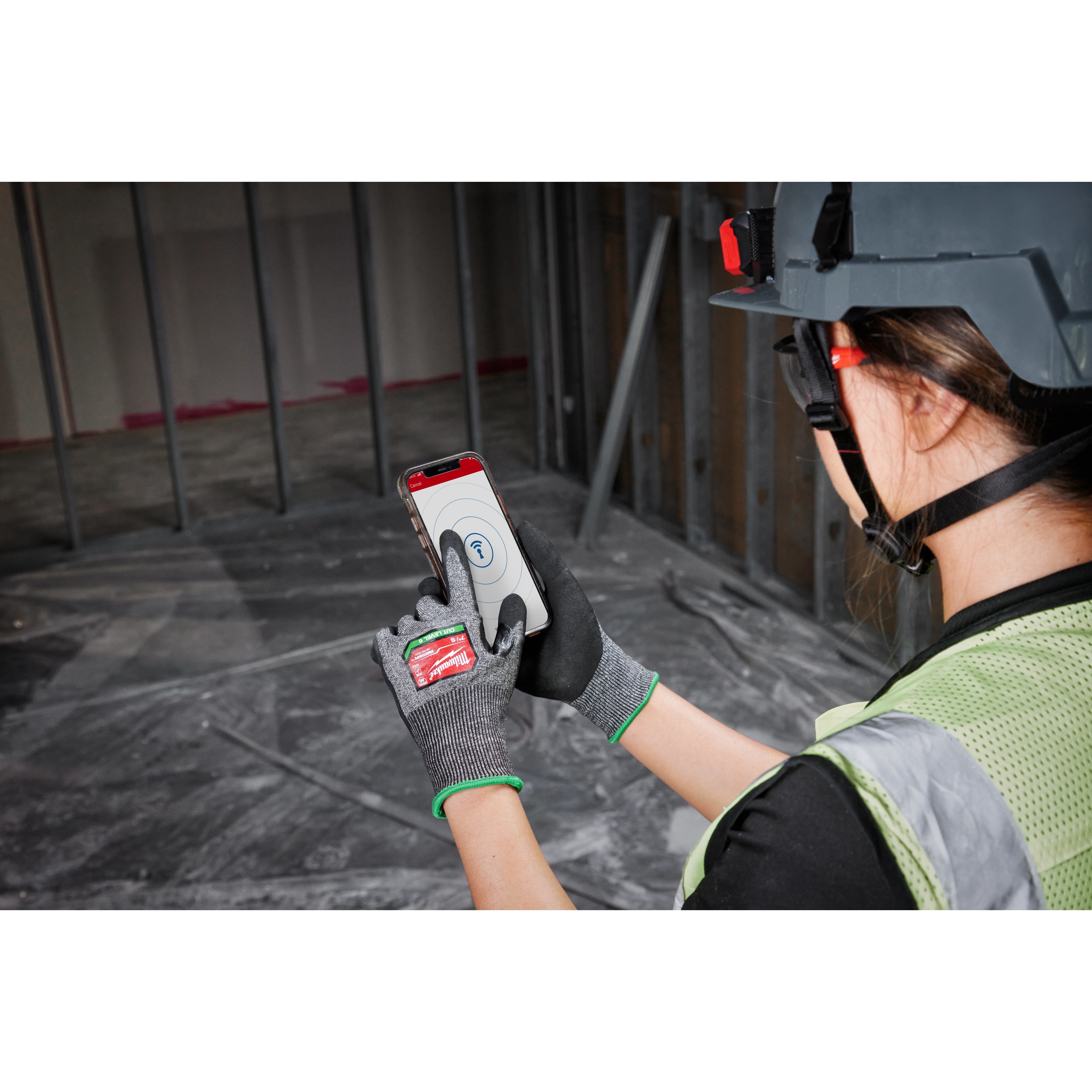 Person wearing Cut Level 6 High-Dexterity Nitrile Dipped Gloves - S, hard hat, and safety vest, using a smartphone on a construction site.