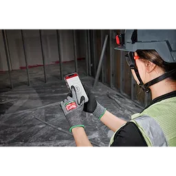 Person wearing Cut Level 6 High-Dexterity Nitrile Dipped Gloves - S, hard hat, and safety vest, using a smartphone on a construction site.