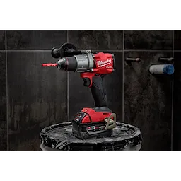 Power drill with a 1/4" Diamond Grit Hole Saw attachment, placed on a paint-splattered stool against a dark tiled wall background.